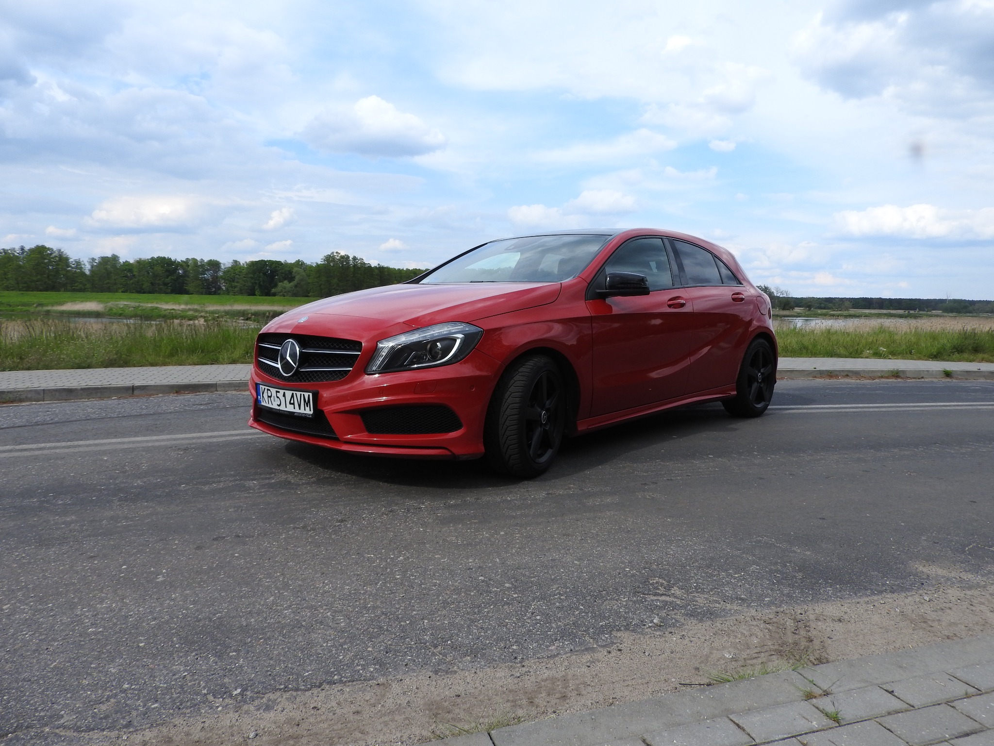 Czerwony Mercedes A Klasa z czarnymi felgami na szarej asfaltowej drodze, w tle zielone drzewa i błękitne niebo z chmurami.