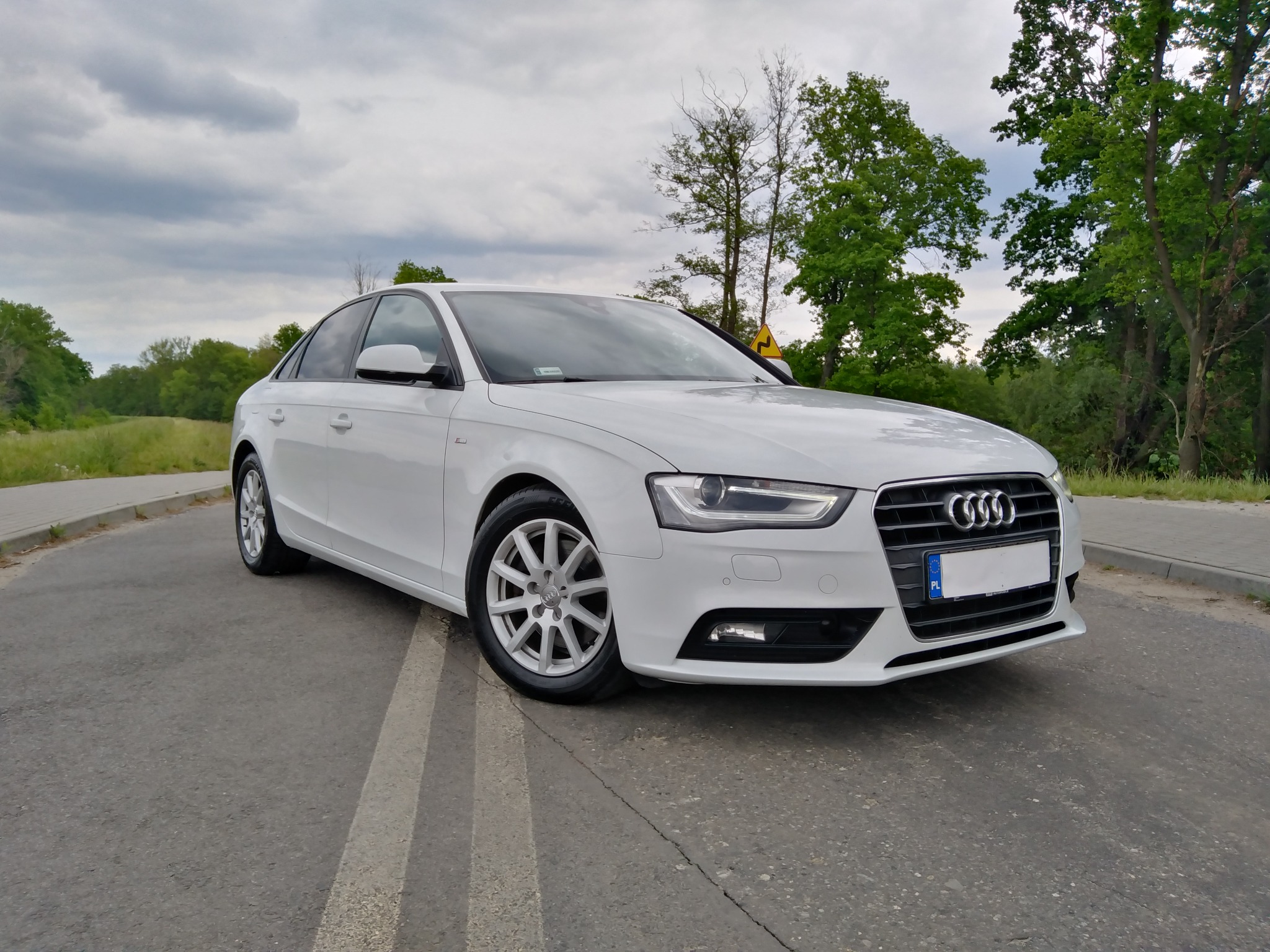 Białe Audi A4 zaparkowane na asfaltowej drodze z widocznym znakiem drogowym w tle, uchwycone w pochmurny dzień.