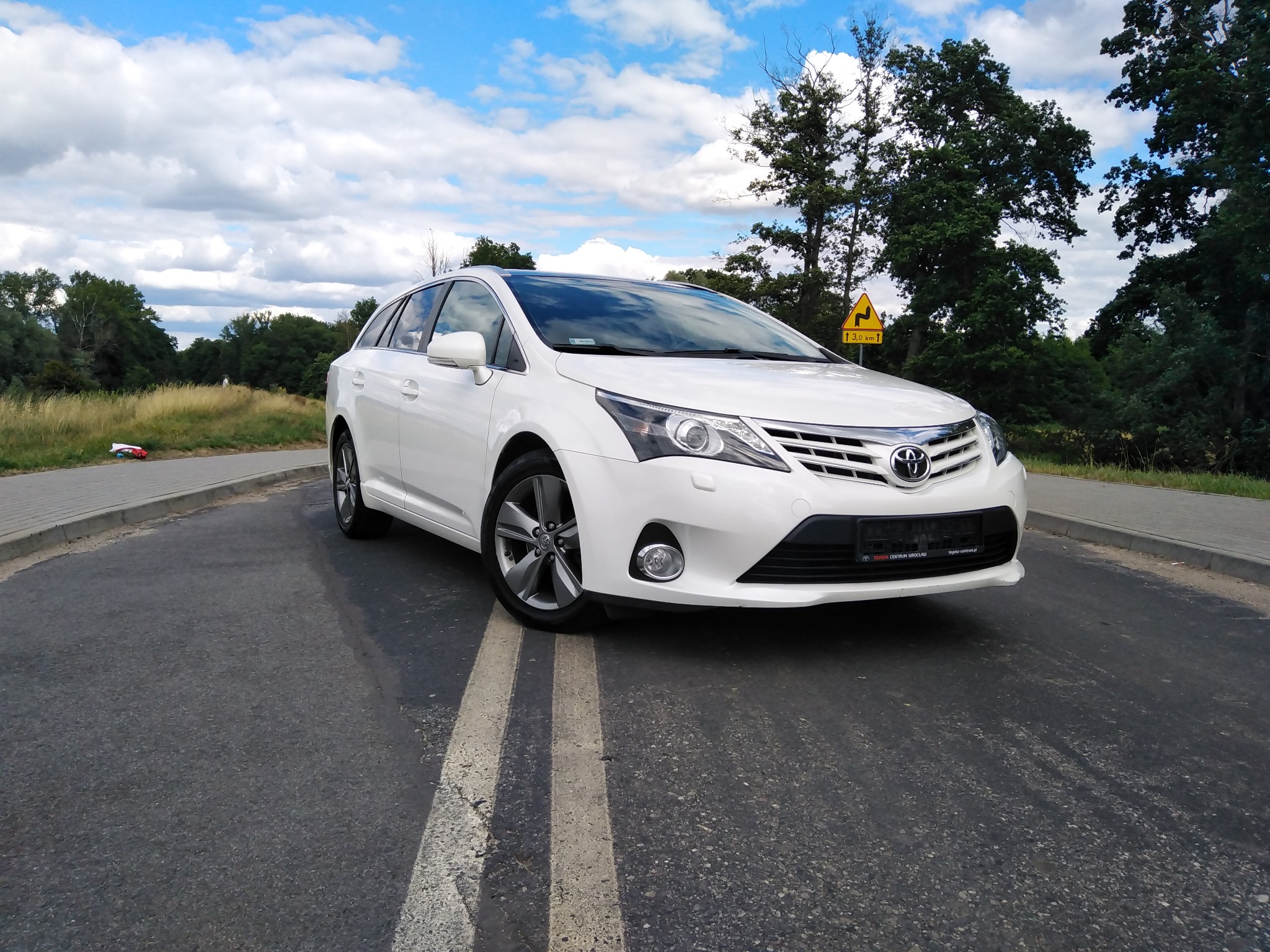 Biała Toyota Avensis kombi zaparkowana na asfalcie z podwójną linią, z widocznym znakiem drogowym i drzewami w tle pod błękitnym niebem z chmurami.