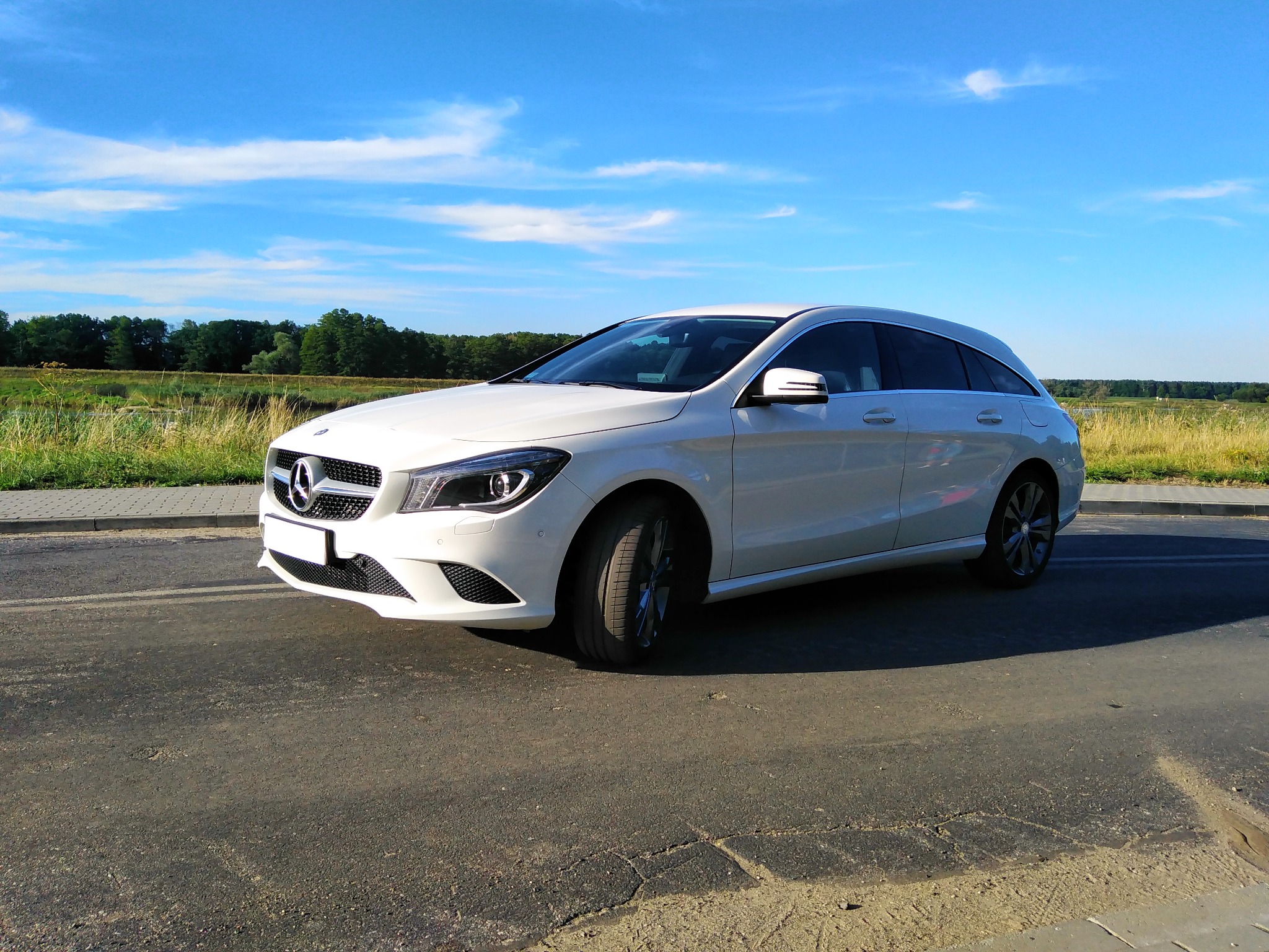 Biały Mercedes CLA Shooting Brake zaparkowany na asfaltowej drodze z widokiem na łąki i błękitne niebo z chmurami w tle.