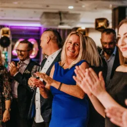 Uśmiechnięta blond kobieta w niebieskiej sukience klaszcze w dłonie podczas imprezy z balonami w tle i innymi gośćmi.