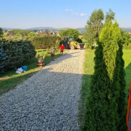 Żwirowa droga w trakcie budowy, z widocznym mini-walcem, dwoma pracownikami i koparką w tle, otoczona zielenią i drzewami w słoneczny dzień.