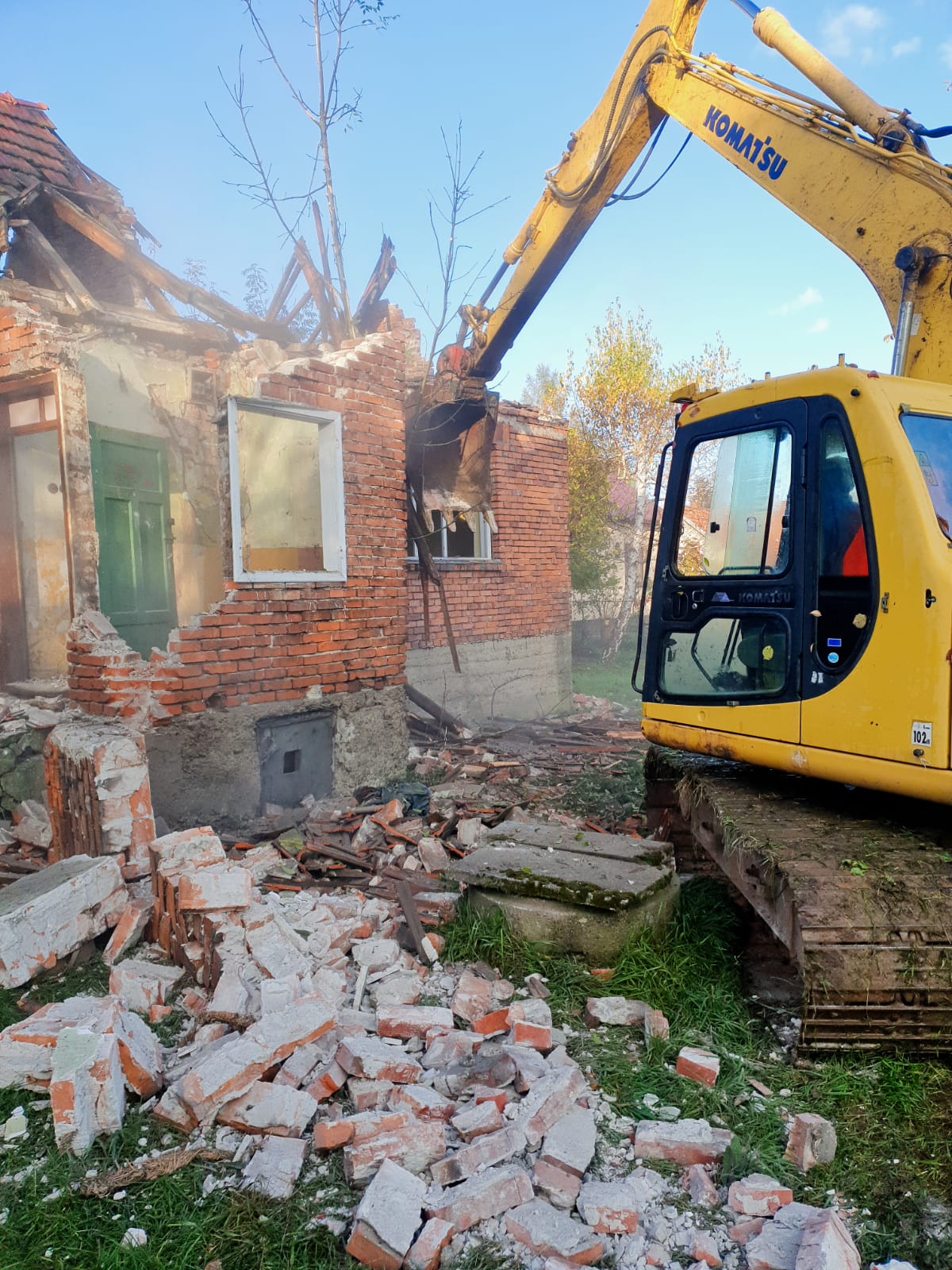 Żółta koparka Komatsu w trakcie burzenia ceglanego domu z zielonymi drzwiami; widoczne gruzy i fragmenty ścian, niebo w tle.
