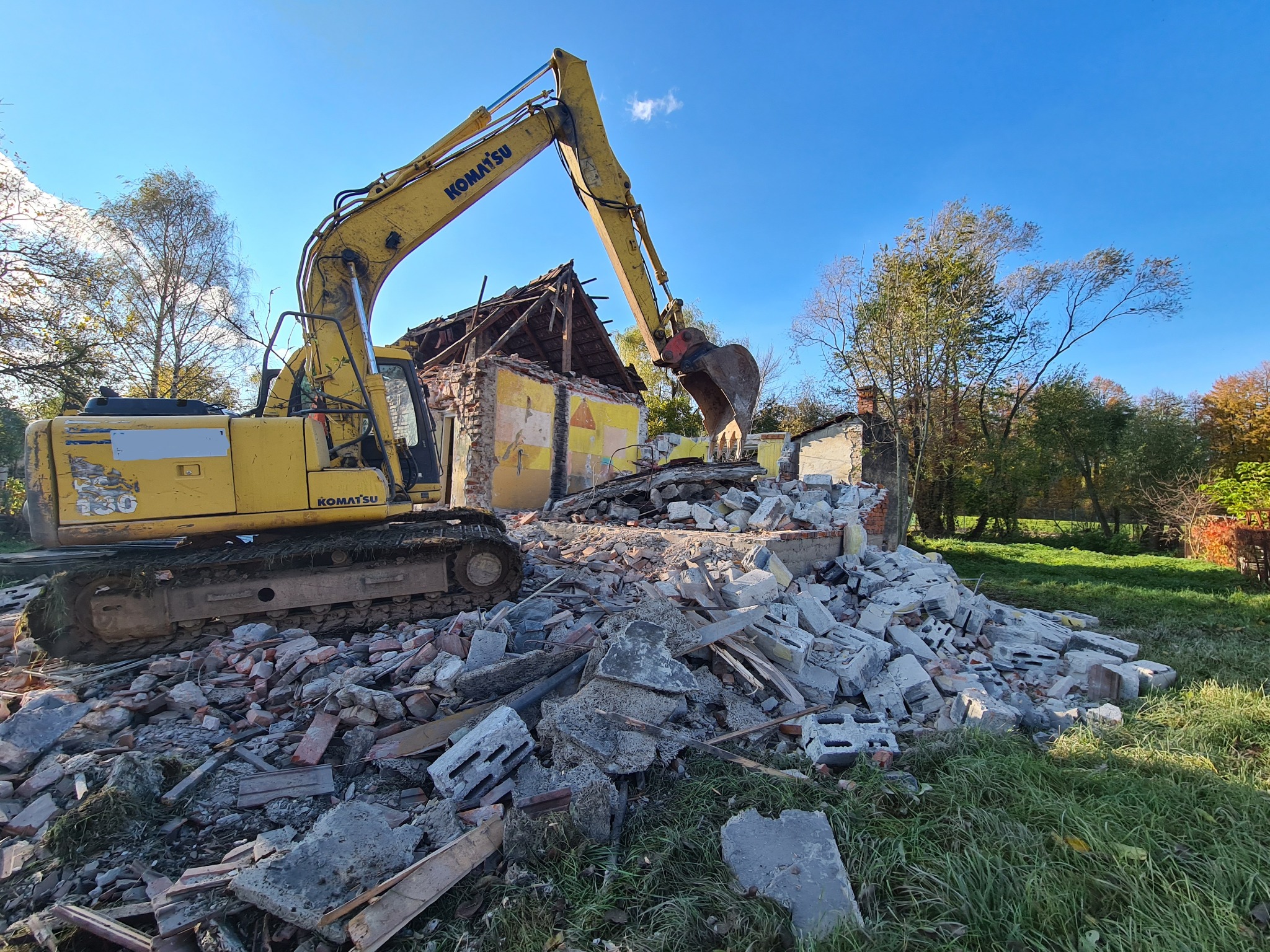 Żółta koparka Komatsu demontuje częściowo zburzony budynek z cegły i pustaków na tle błękitnego nieba i drzew