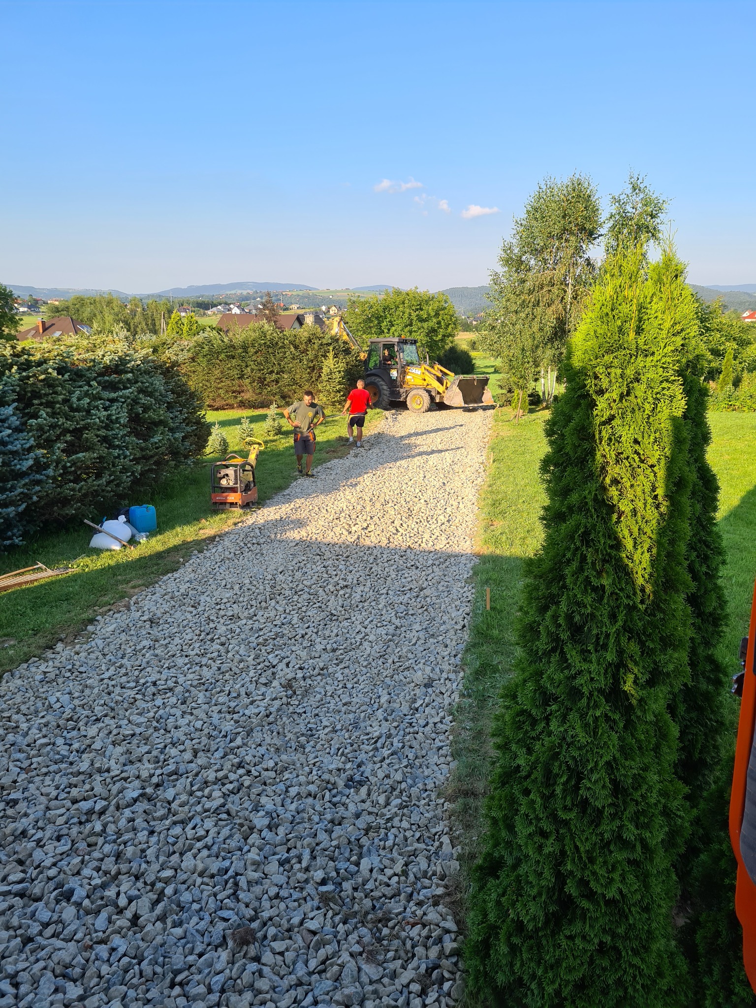 Żwirowa droga w trakcie budowy, z widocznym mini-walcem, dwoma pracownikami i koparką w tle, otoczona zielenią i drzewami w słoneczny dzień.