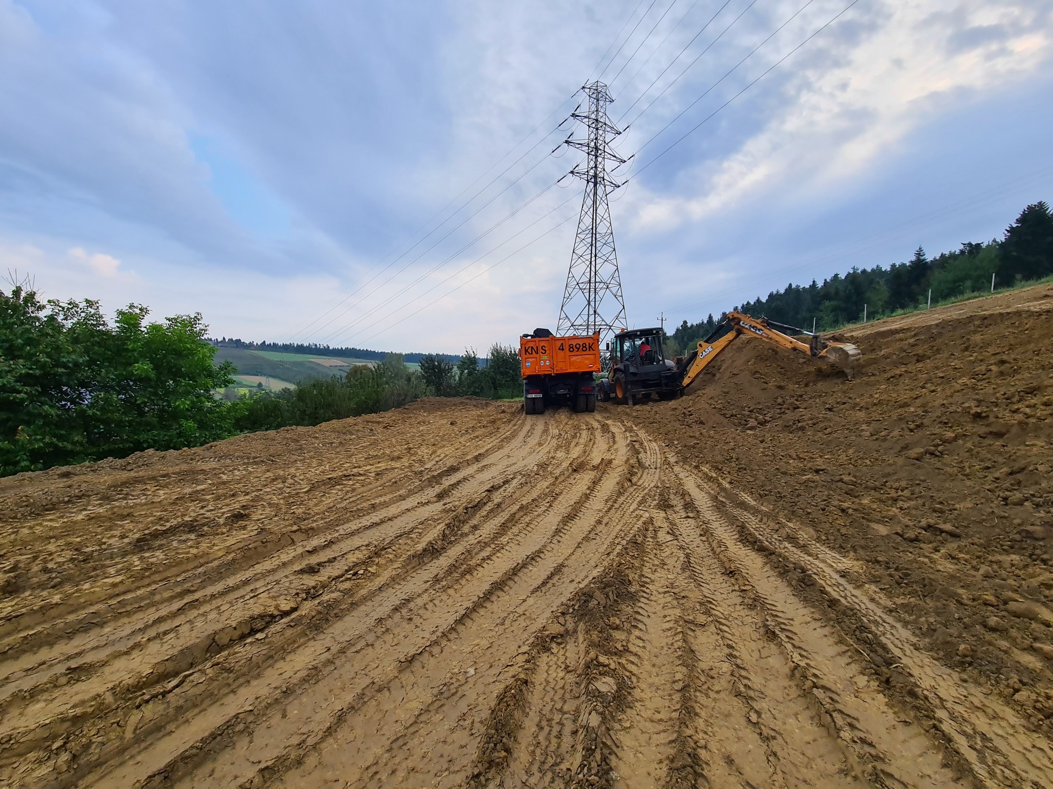 Prace ziemne w Nowym Sączu: pomarańczowa wywrotka i koparko-ładowarka CASE wyrównują teren pod linią wysokiego napięcia. Widoczne głębokie ślady opon w gruncie.