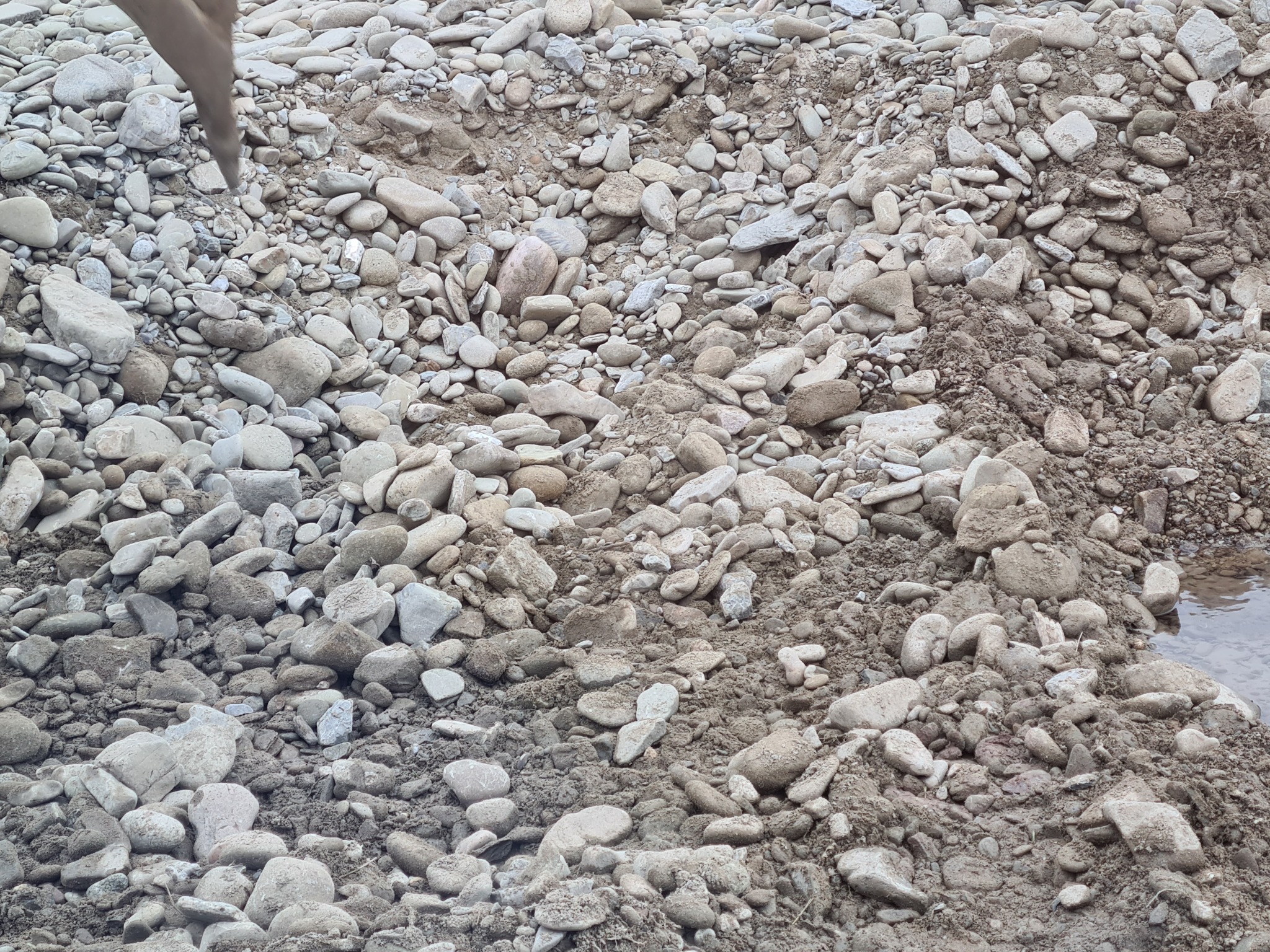Stos kamieni i żwiru o różnej wielkości, częściowo pokrytych błotem, z niewielkim fragmentem wody widocznym w prawym dolnym rogu.