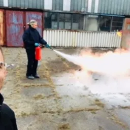 Mężczyzna robiący selfie na tle osoby w kombinezonie gaszącej ogień gaśnicą proszkową na placu manewrowym z kontenerami w tle.