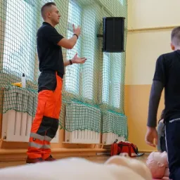 Instruktor w czarnej koszulce i pomarańczowych spodniach demonstruje techniki ratownicze na manekinie przed grupą uczniów w sali gimnastycznej.