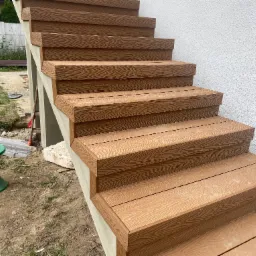 Schody zewnętrzne wykończone deską kompozytową o fakturze drewna, prowadzące do budynku z białą elewacją. Widoczny fragment ogrodu z zielonym stołkiem.