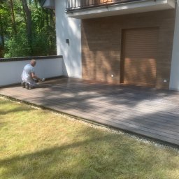PHU Malbis Leszek Kaleta - Pracownik w trakcie olejowania drewnianego tarasu przy nowoczesnym domu z elementami drewnianej elewacji i zielonym trawnikiem. Widoczne narzędzia i etap impregnacji.