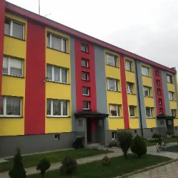 Wielobarwny budynek wielorodzinny po renowacji elewacji: żółte, czerwone i szare pasy. Widoczne okna, wejścia i otoczenie z zielenią.