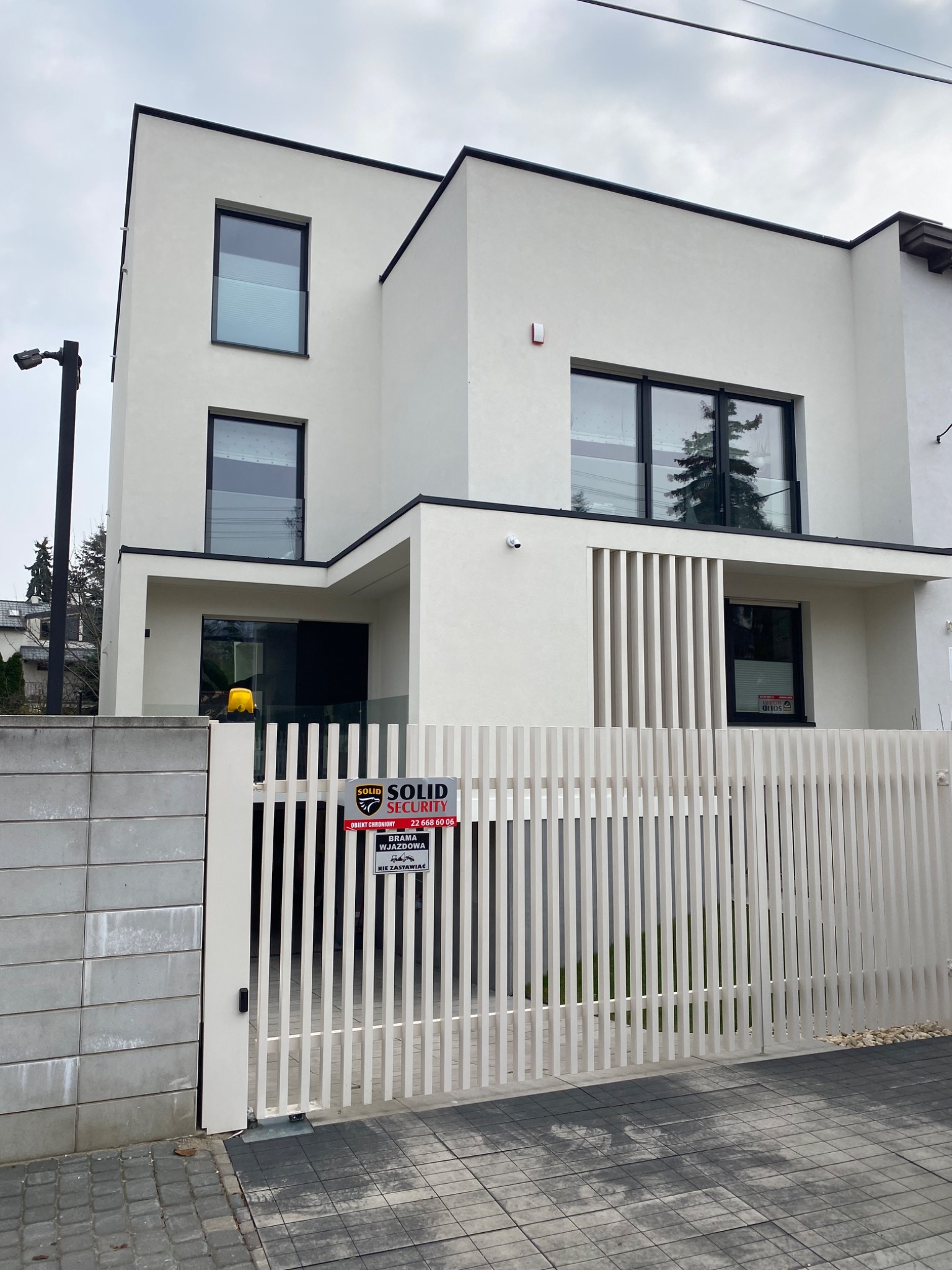 Nowoczesny, biały dom z czarnymi ramami okiennymi, widziany zza białego ogrodzenia z pionowych desek. Na ogrodzeniu tabliczka firmy ochroniarskiej.