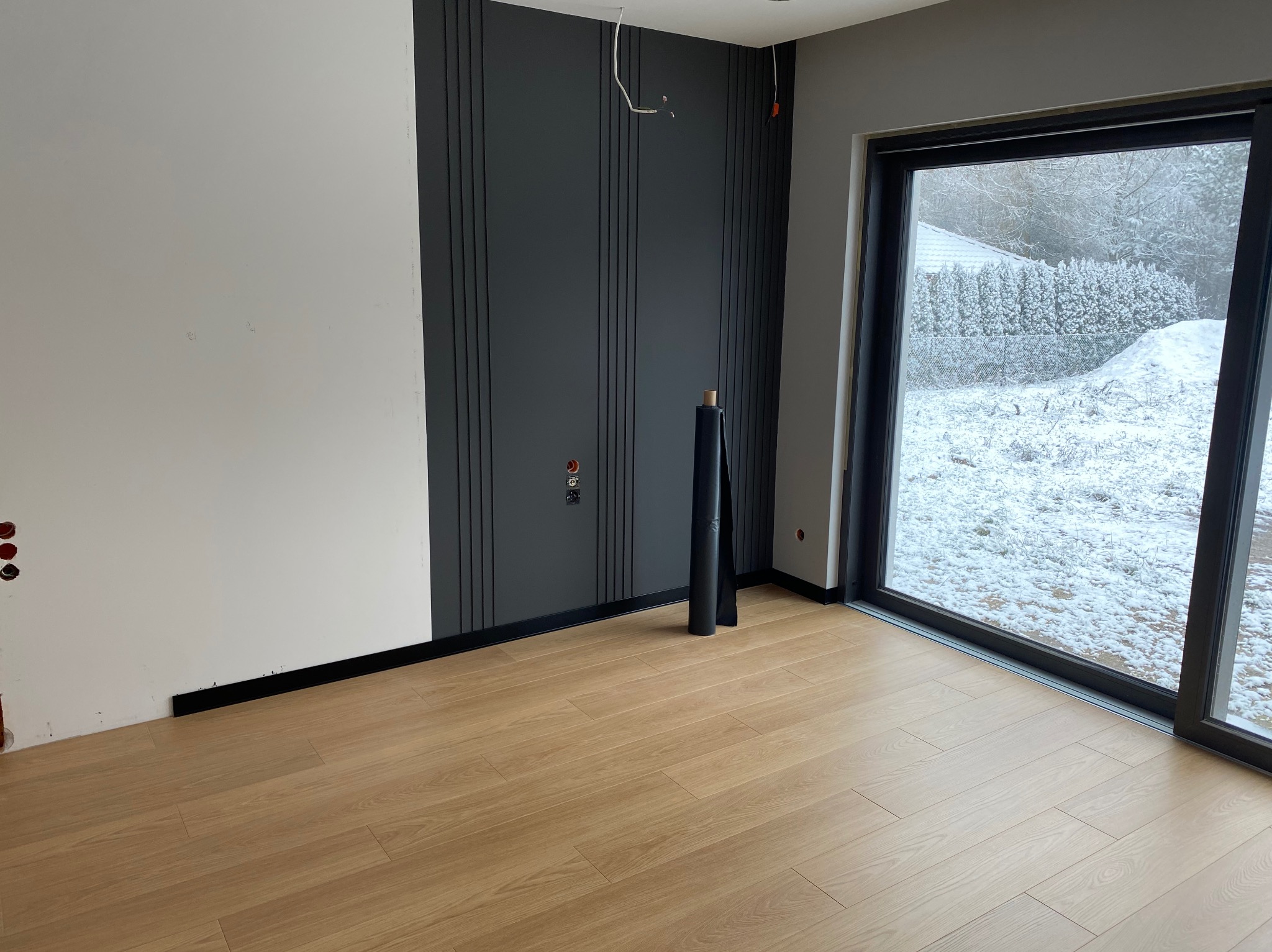 Wnętrze w trakcie wykańczania: panele ścienne z listwami, okno z widokiem na zimowy krajobraz, fragment podłogi i ściany przygotowane do dalszych prac.