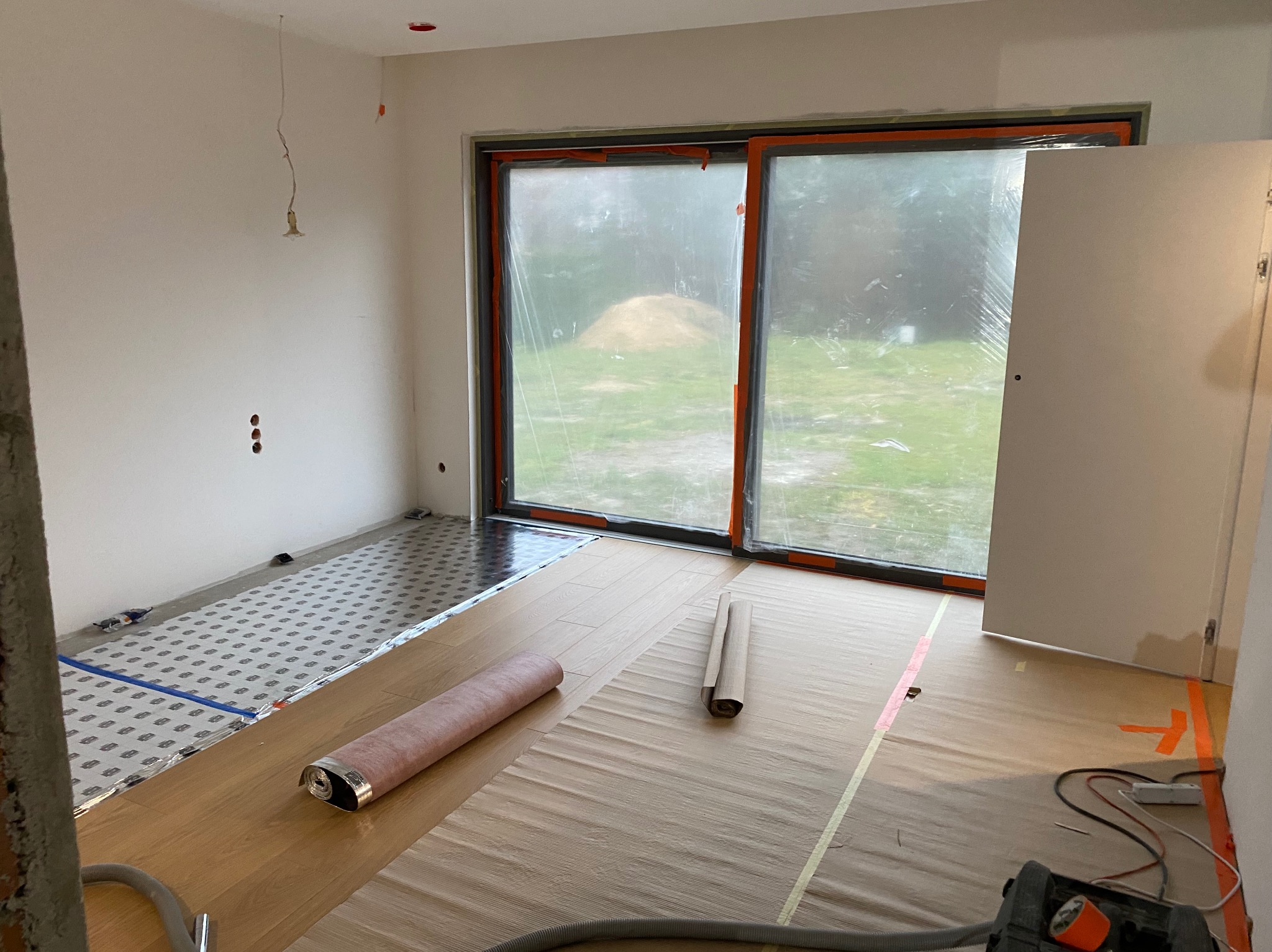 Wnętrze w trakcie wykończenia: podkład pod panele podłogowe, okna zabezpieczone folią, widoczne przygotowania do dalszych prac wykończeniowych. Surowy stan.