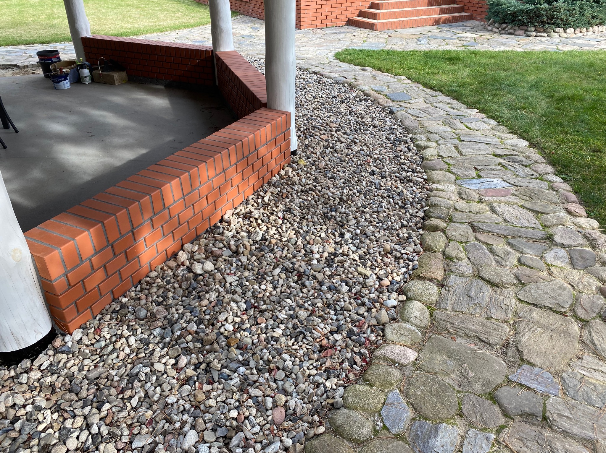 Ceglany murek oddzielający żwirowe podłoże od kamiennej ścieżki w ogrodzie. Widoczne białe kolumny i fragment budynku z cegły.