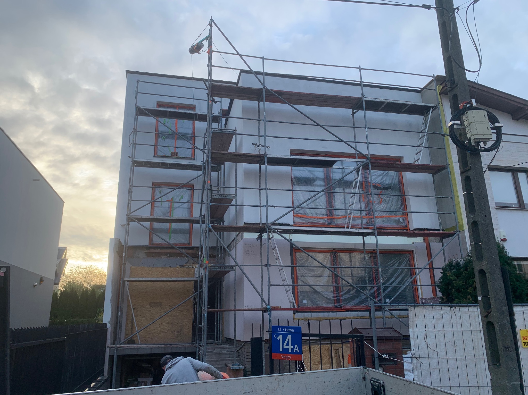 Dom w trakcie ocieplania: rusztowania, okna zabezpieczone folią, widoczny fragment elewacji z ociepleniem. Prace budowlane w toku, tabliczka z adresem.