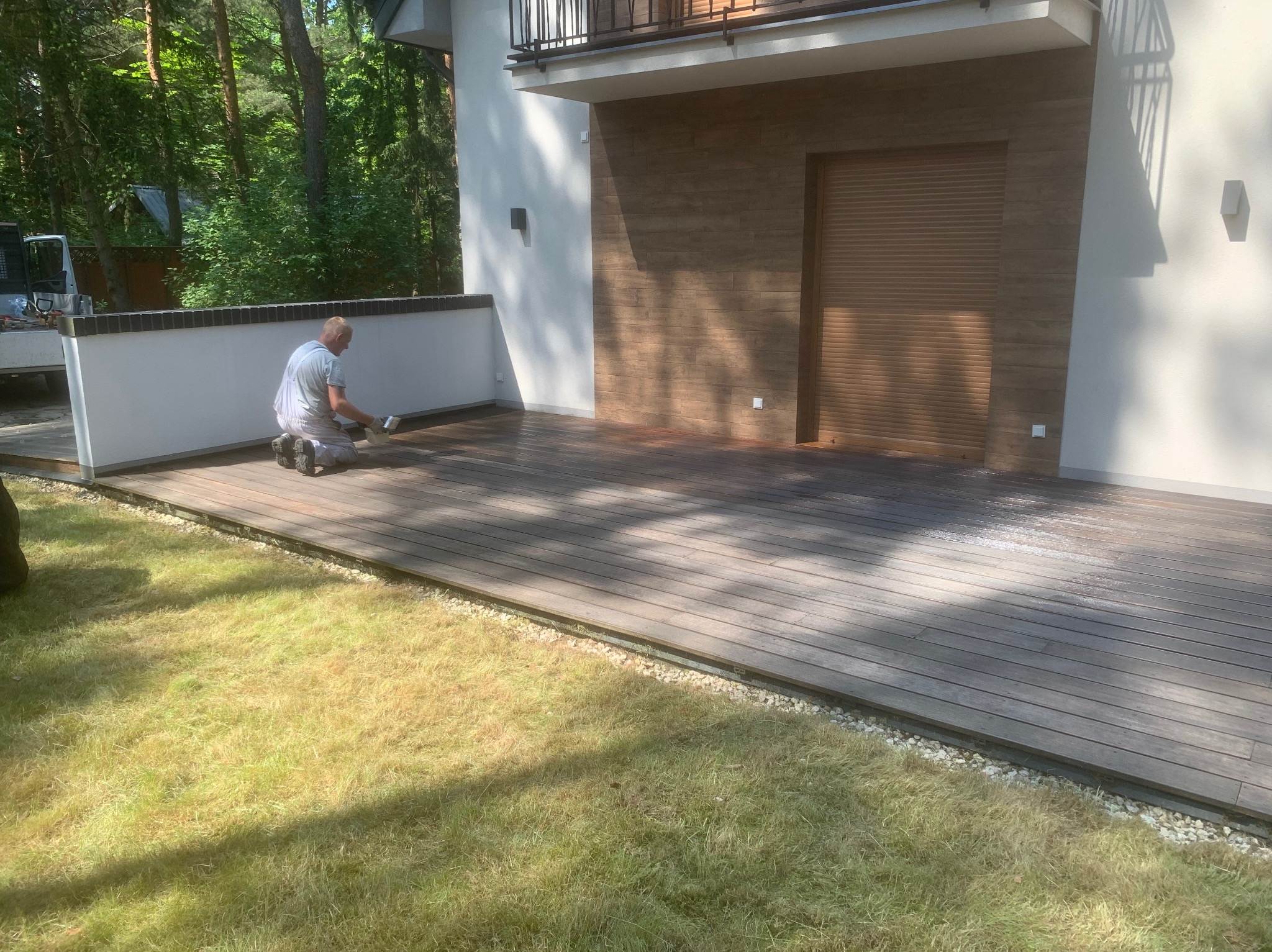 Pracownik w trakcie olejowania drewnianego tarasu przy nowoczesnym domu z elementami drewnianej elewacji i zielonym trawnikiem. Widoczne narzędzia i etap impregnacji.