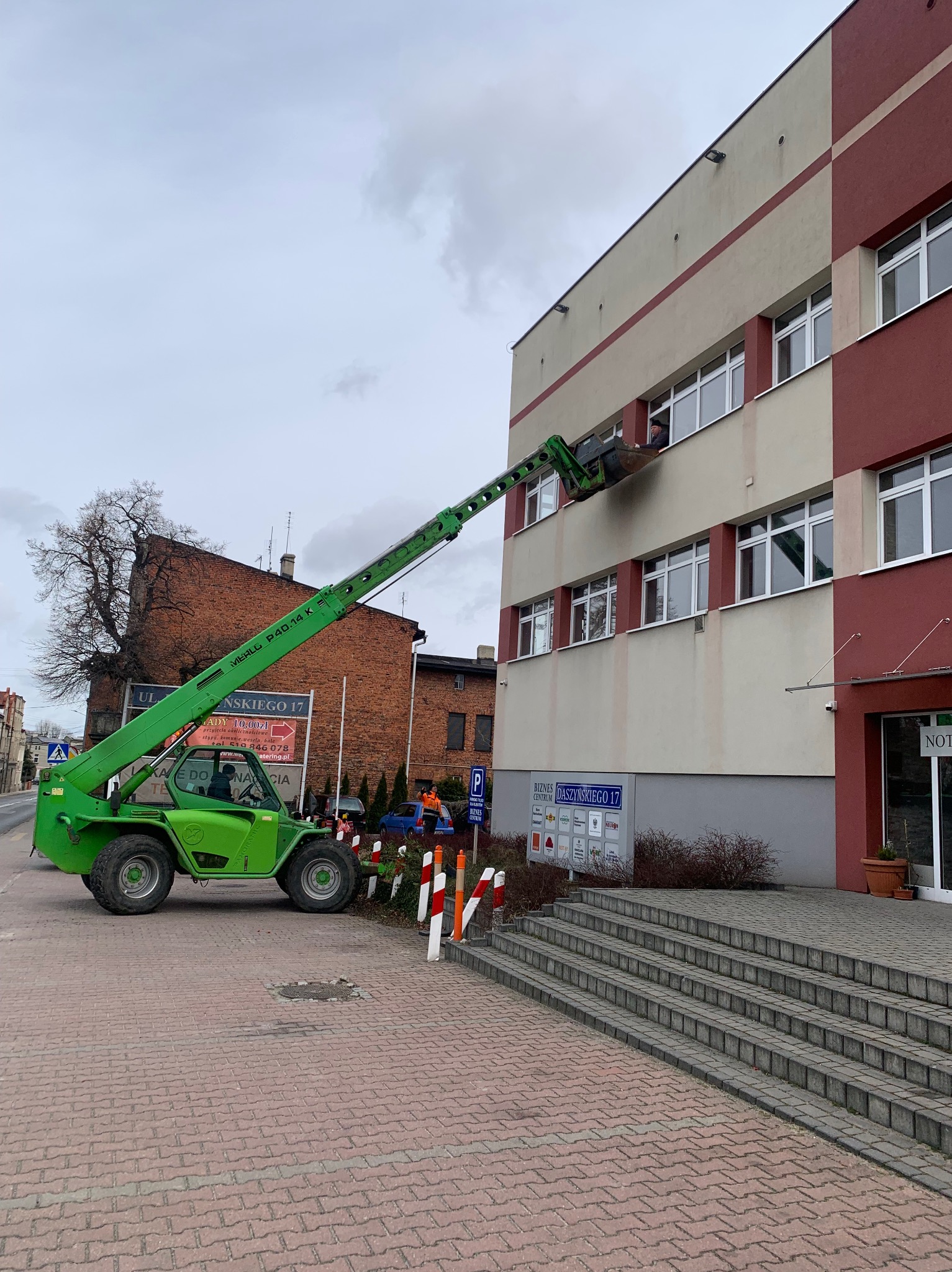 Prace na wysokościach przy budynku z użyciem podnośnika koszowego Merlo P40.14 K. Widoczny pracownik w koszu oraz elewacja budynku w odcieniach beżu i czerwieni.