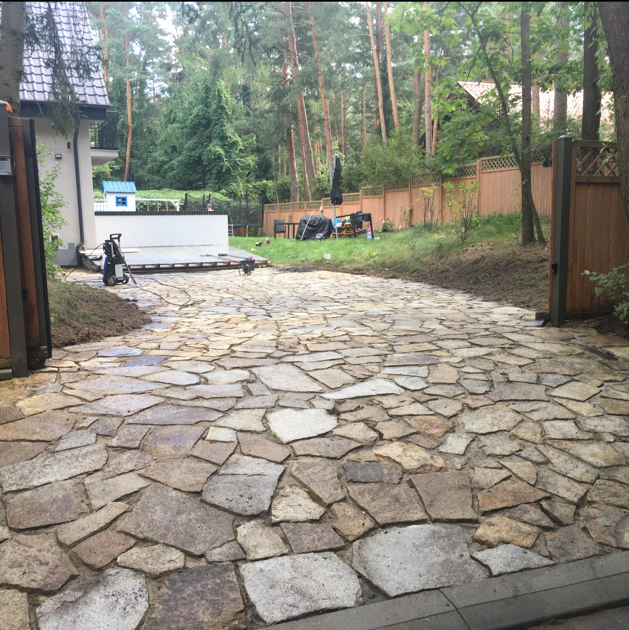 Ułożony z nieregularnych kamieni podjazd do domu, widok na posesję z ogrodem i drewnianym ogrodzeniem. W tle zieleń drzew.