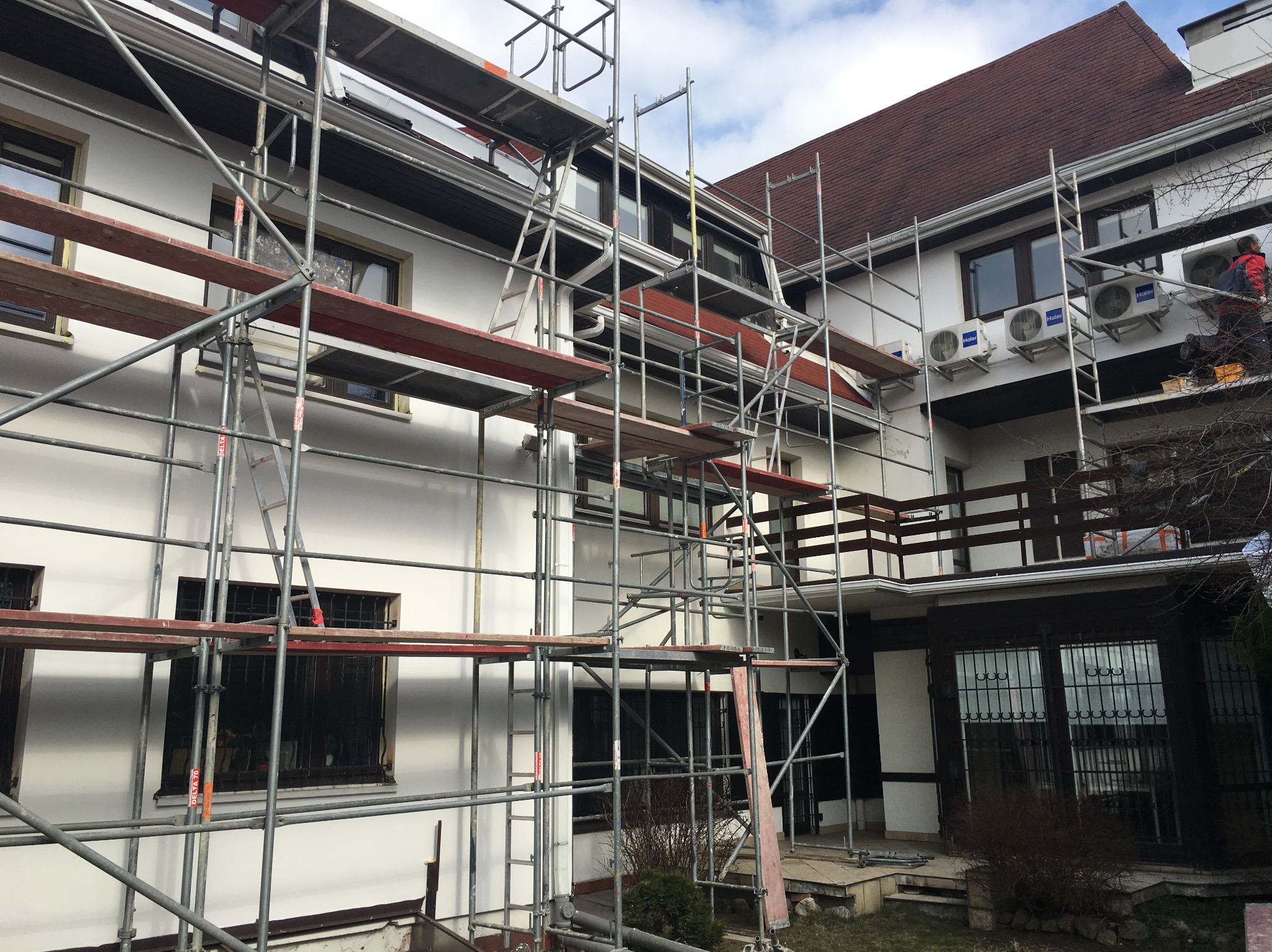 Biały budynek w trakcie remontu elewacji, otoczony rusztowaniami. Widoczny pracownik na rusztowaniu i zamontowane klimatyzatory. Częściowo odsłonięty dach z brązowej dachówki.
