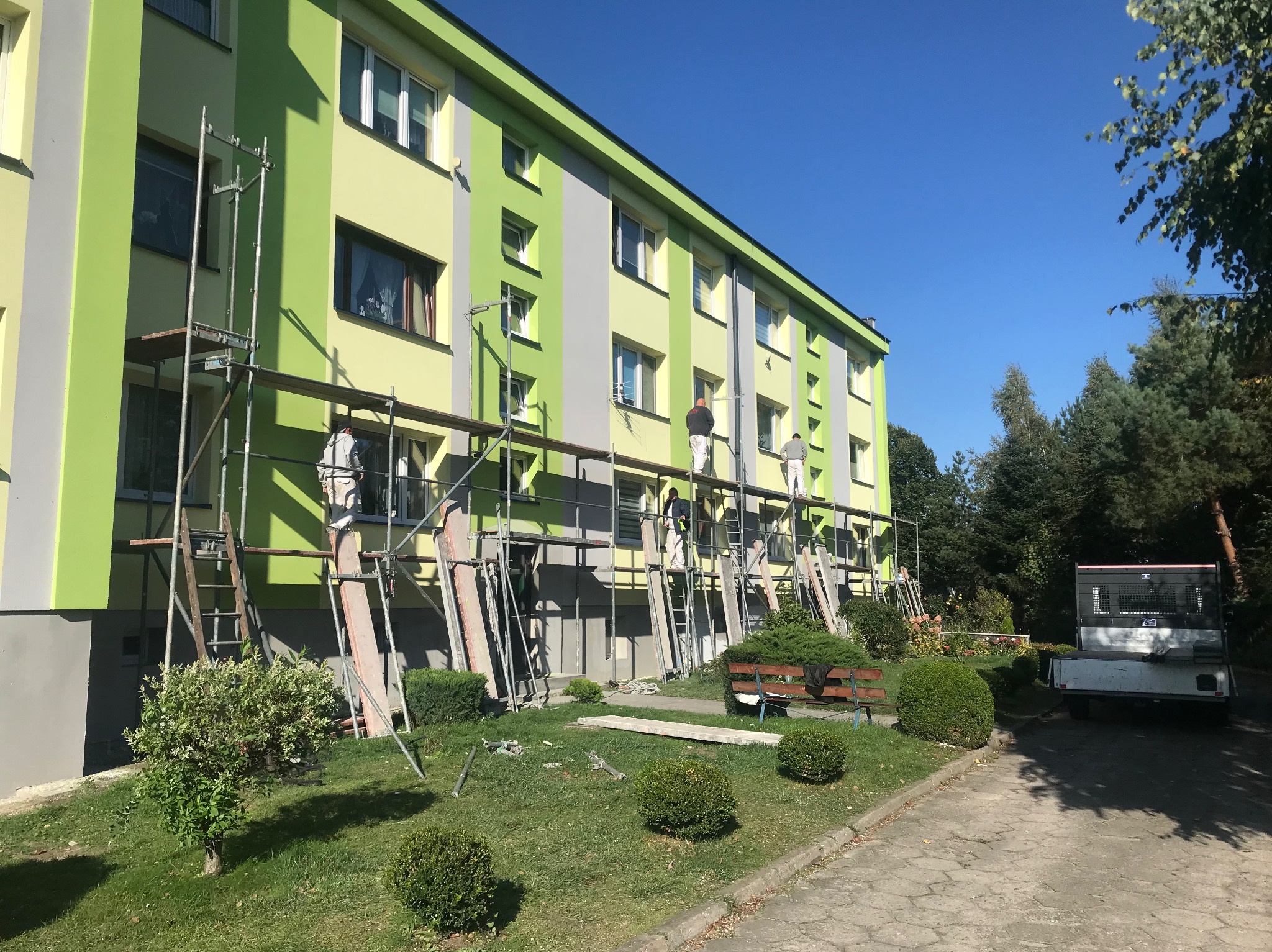 Malowanie elewacji bloku mieszkalnego z użyciem rusztowań. Widoczni pracownicy w trakcie pracy, budynek w odcieniach zieleni i szarości, błękitne niebo w tle.