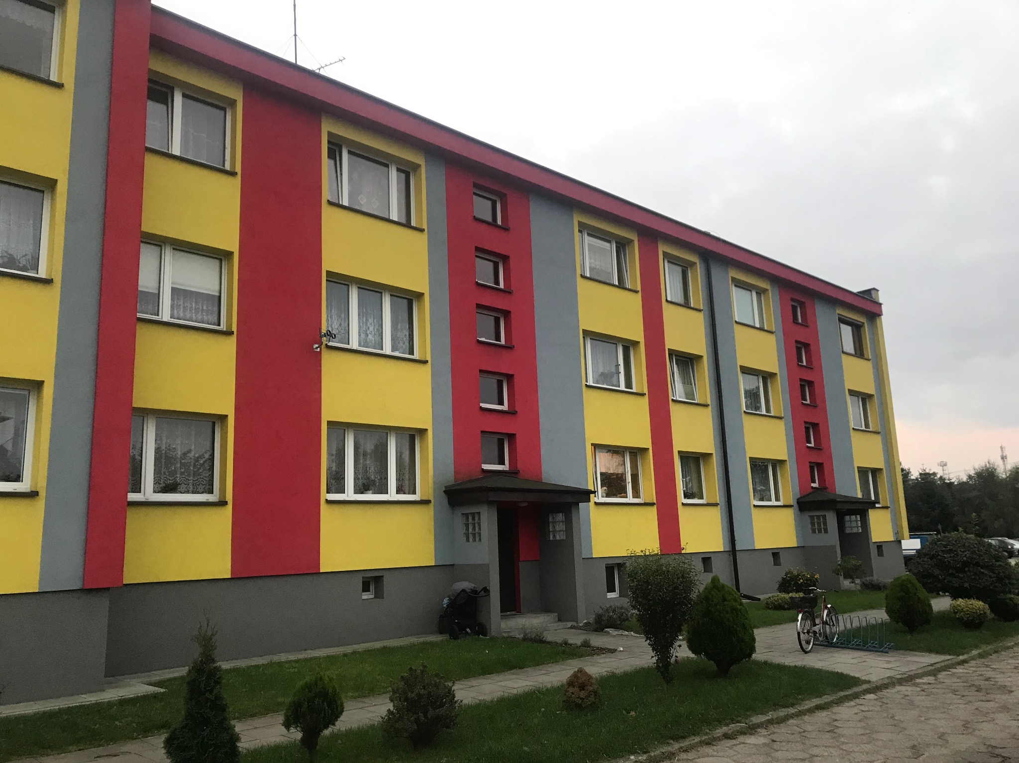 Wielobarwny budynek wielorodzinny po renowacji elewacji: żółte, czerwone i szare pasy. Widoczne okna, wejścia i otoczenie z zielenią.