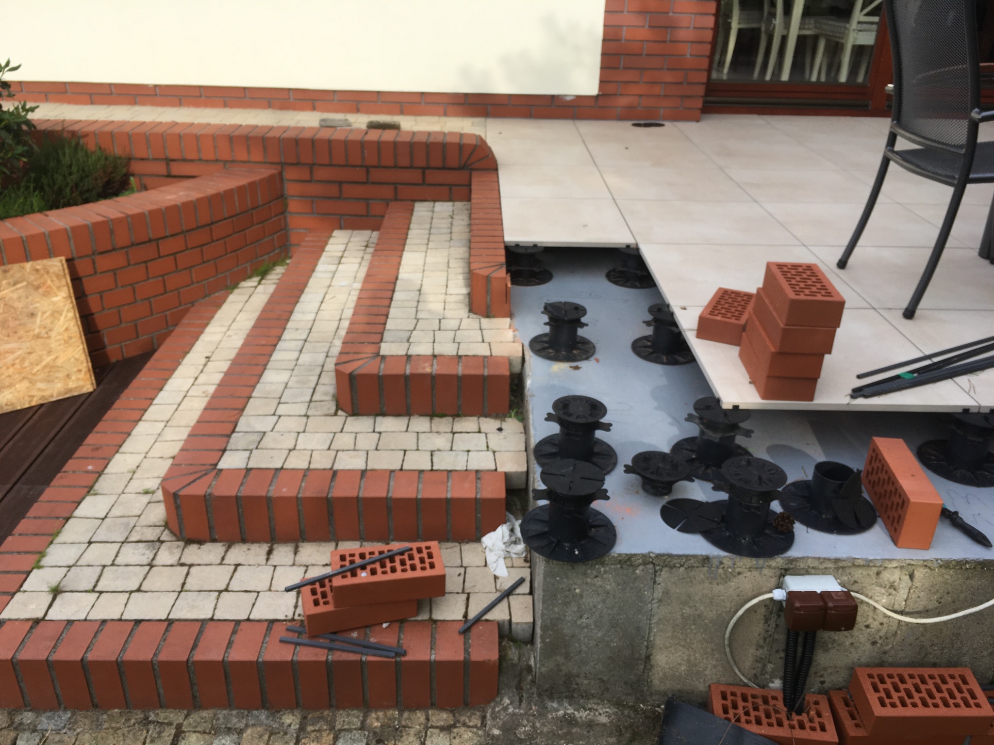 Tarasy wentylowane na regulowanych wspornikach. Wykończenie schodów i obrzeży czerwoną cegłą klinkierową. Częściowo ułożona podłoga tarasu.