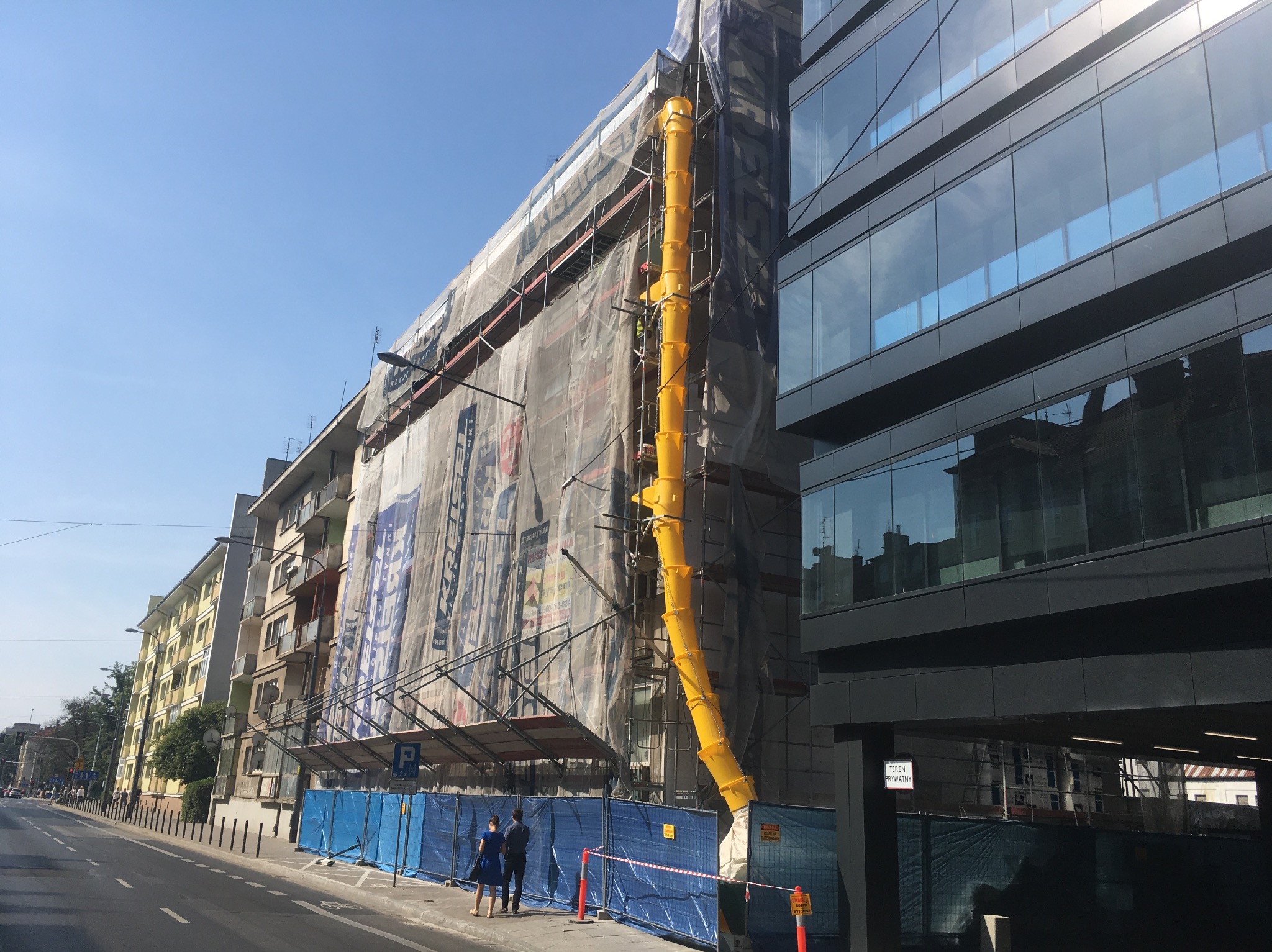 Budynek w rusztowaniach, osłonięty siatką, z żółtym zsypem na gruz. Dwie osoby stoją na chodniku przed budynkiem. Widok na ulicę i sąsiedni nowoczesny budynek.