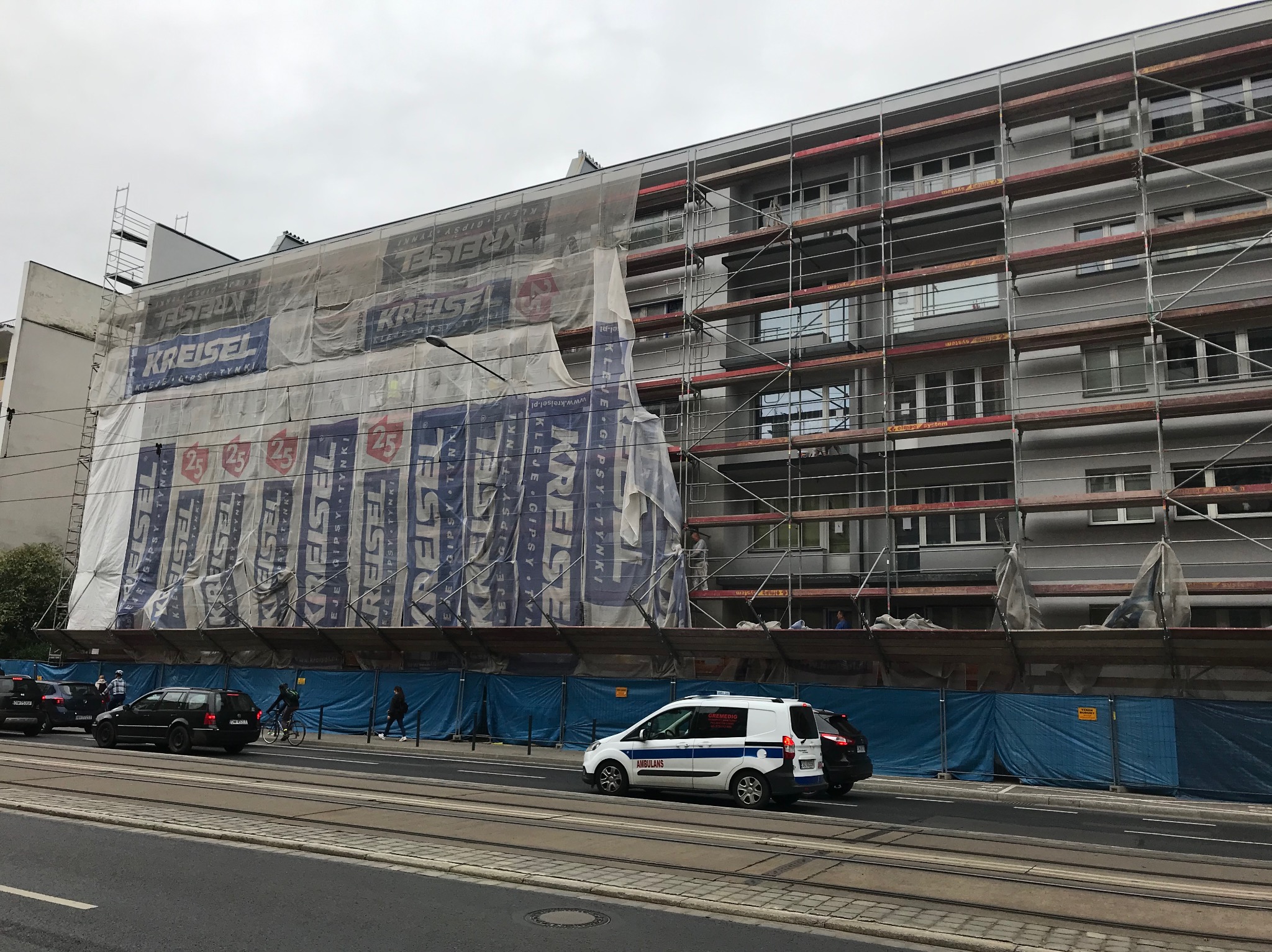 Ocieplanie bloku mieszkalnego w rusztowaniach, reklama Kreisel, przysłonięta siatką. Na dole ulica, pojazdy, w tym ambulans. Pochmurny dzień.