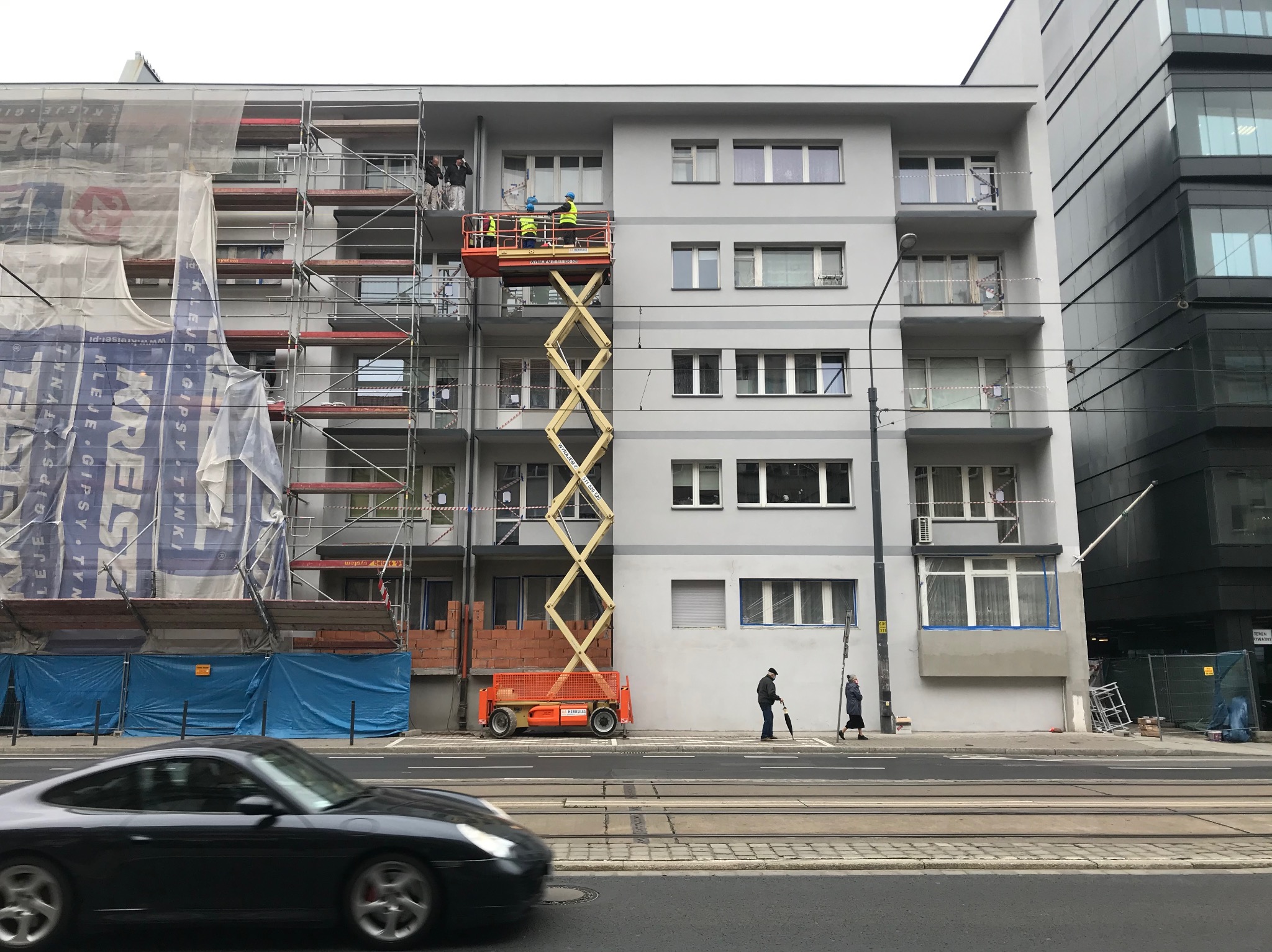 Elewacja budynku w trakcie remontu: rusztowania, podnośnik z pracownikami w żółtych kamizelkach. Przechodnie na chodniku i przejeżdżający samochód. Fragment budynku w siatce.