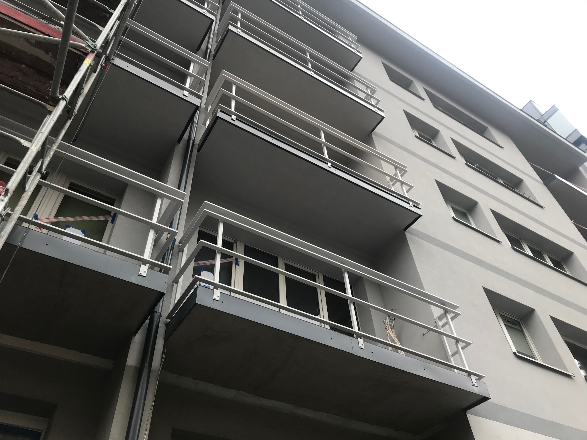 Elewacja budynku z balkonami w trakcie prac wykończeniowych; widoczne rusztowania i zabezpieczenia. Ujęcie z dołu, szara kolorystyka.