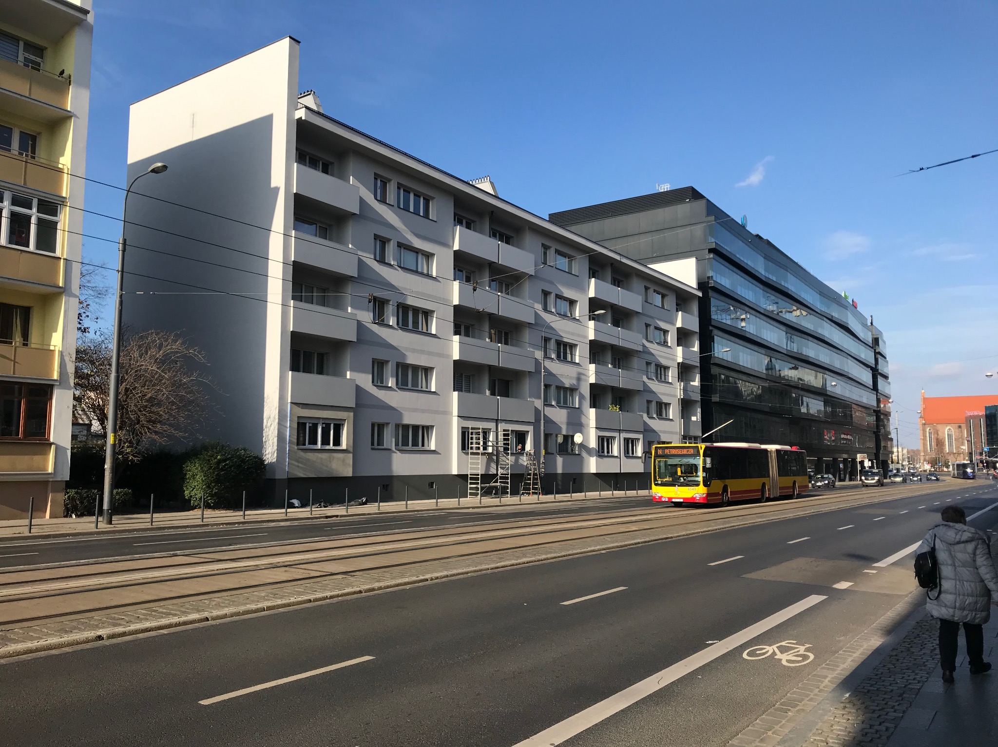 Odnowiona elewacja bloku z balkonami w miejskiej scenerii. Na parterze widoczne rusztowania, sygnalizujące trwające prace. W tle nowoczesny biurowiec i tramwaj.