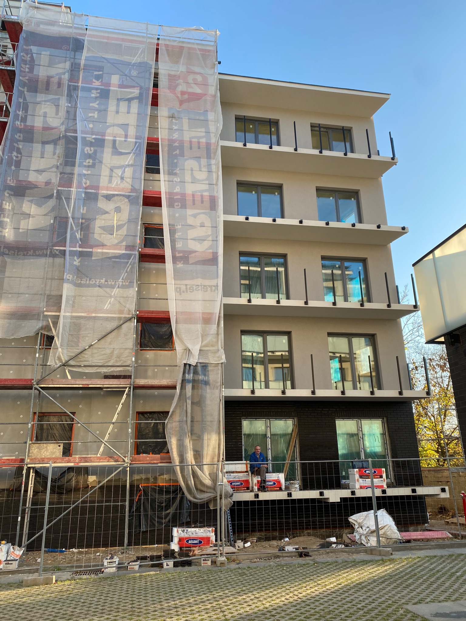 Budynek w trakcie ocieplania, rusztowania z siatką ochronną, widoczna warstwa izolacyjna. Część elewacji już wykończona, z oknami i balkonami. Prace w toku.