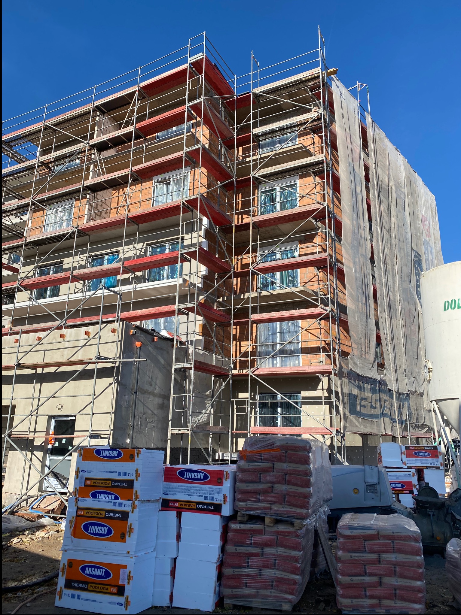 Budynek w trakcie ocieplania, otoczony rusztowaniami. Widoczne materiały izolacyjne i worki z zaprawą. Część elewacji zakryta siatką ochronną. Słoneczny dzień.