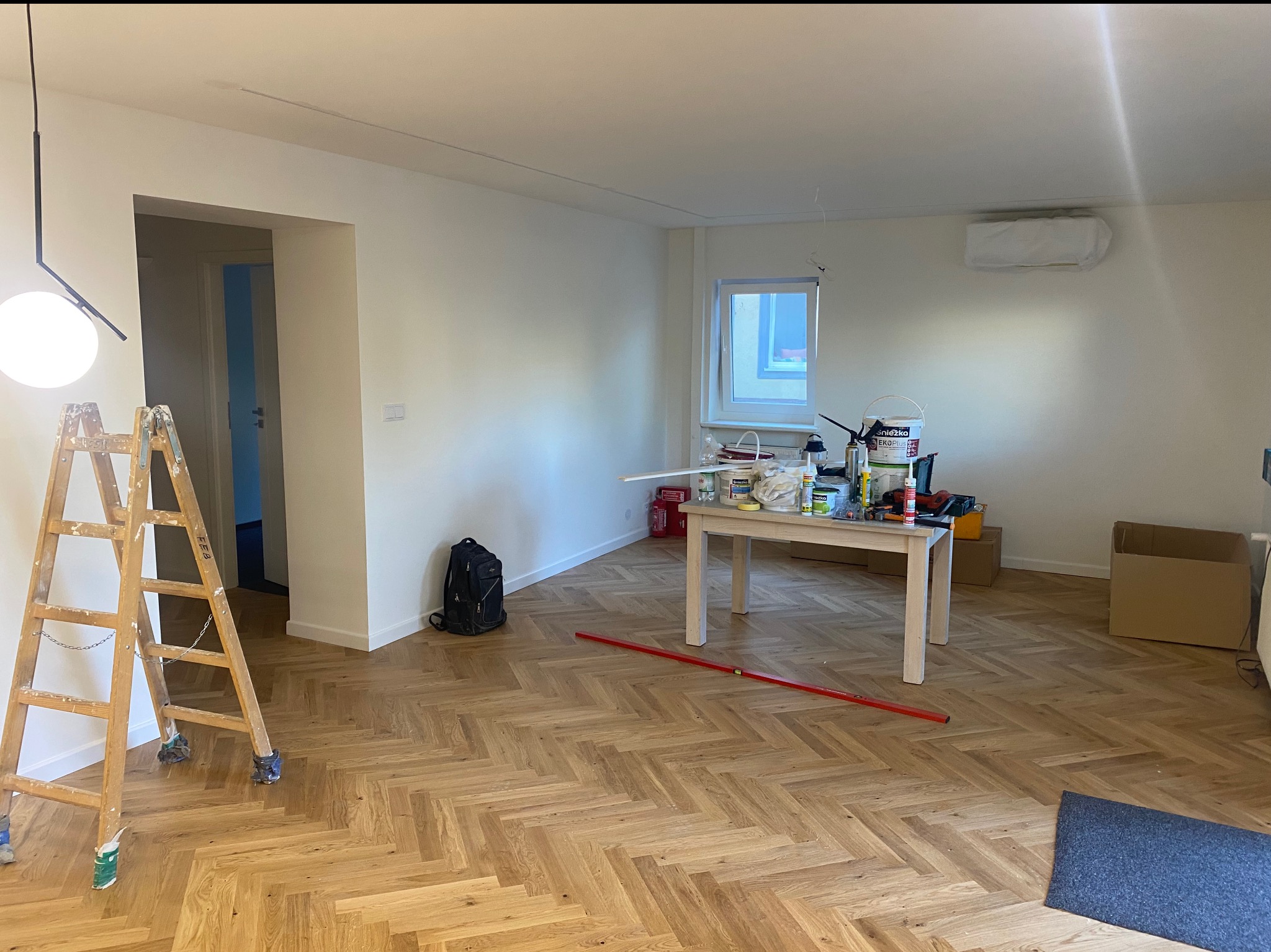 Jasne wnętrze w trakcie prac wykończeniowych: parkiet w jodełkę, drabina malarska, stół z narzędziami i farbami. Widoczny fragment sufitu z klimatyzacją.