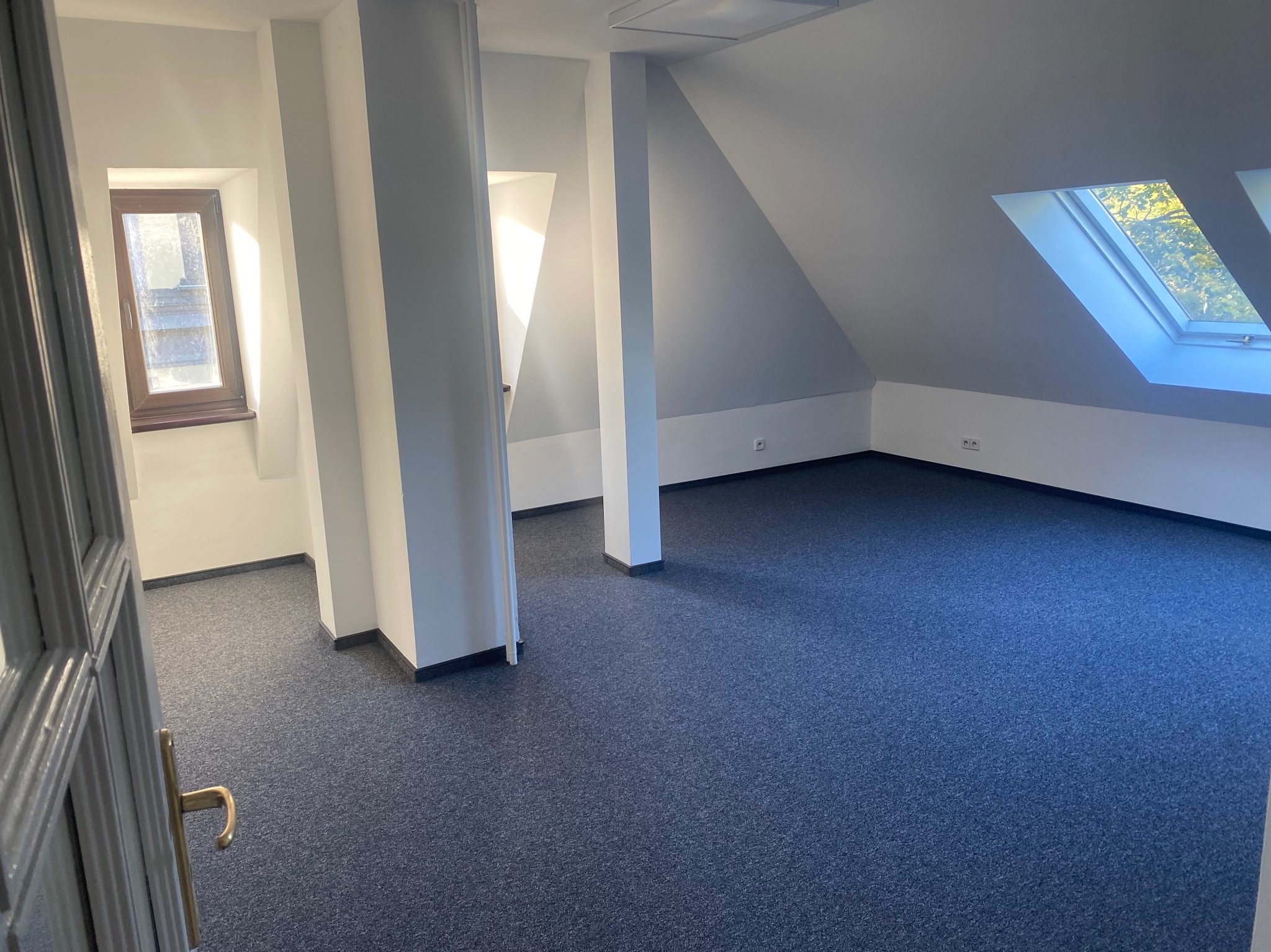 Poddasze biurowe po remoncie: białe ściany, kolumny, okna dachowe i wykładzina w odcieniach granatu. Widok zza uchylonych drzwi.