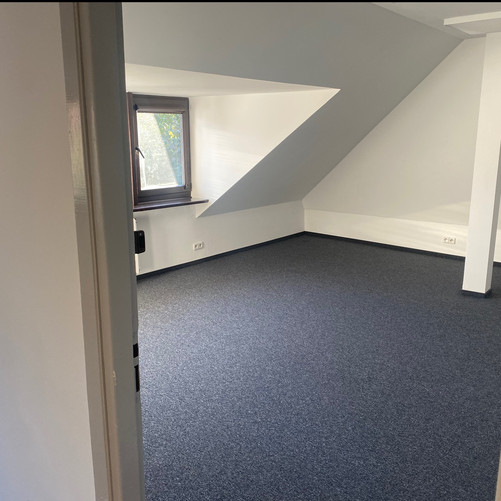 Poddasze po remoncie: szara wykładzina, białe ściany, okno w skosie dachu. Widok zza otwartych drzwi. Gniazdka elektryczne przy podłodze.