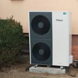 Biała pompa ciepła Vaillant aroTHERM na betonowym fundamencie obok budynku z krzewem, widoczne przewody.