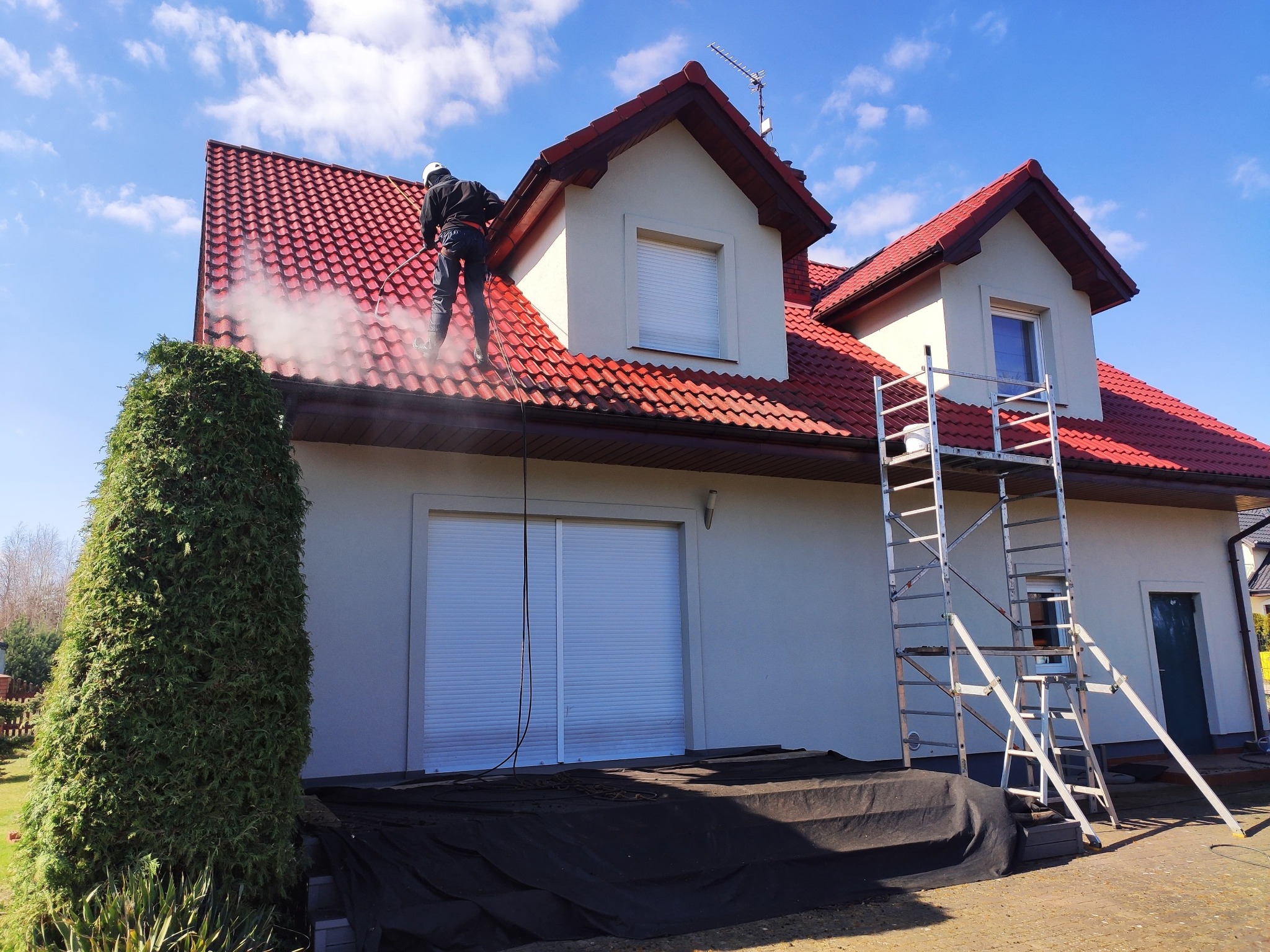 Pracownik w kasku i uprzęży na dachu dwupiętrowego domu z czerwoną dachówką, czyści go za pomocą myjki ciśnieniowej, widoczna para wodna, obok domu rozstawione rusztowanie.