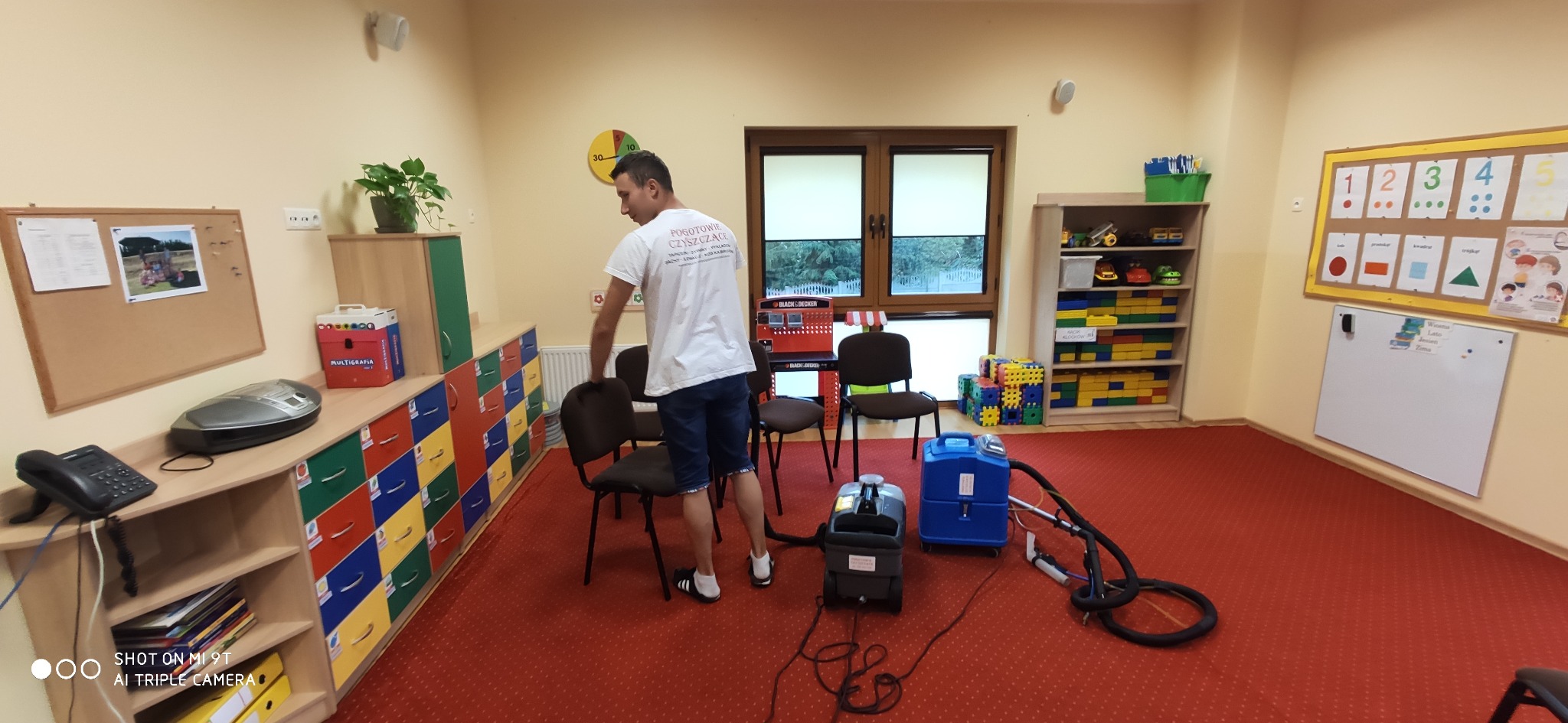 Pracownik firmy sprzątającej przesuwa krzesło, przygotowując się do czyszczenia dywanu w przedszkolu, widoczne dwa odkurzacze piorące.
