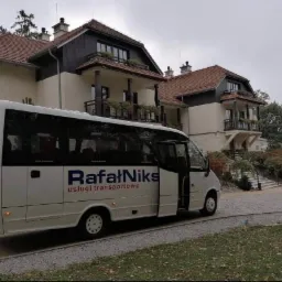 Biały bus z otwartymi tylnymi drzwiami i napisem 'Rafał Niks usługi transportowe' zaparkowany przed budynkiem z balkonami i brązowym dachem, jesienna sceneria.