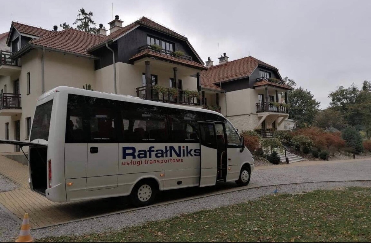 Biały bus z otwartymi tylnymi drzwiami i napisem 'Rafał Niks usługi transportowe' zaparkowany przed budynkiem z balkonami i brązowym dachem, jesienna sceneria.