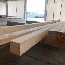 Świeżo obrobione, jasne belki drewniane z czerwonymi oznaczeniami leżące na podłodze w przestronnym magazynie z otwartymi wrotami i widokiem na zewnątrz.