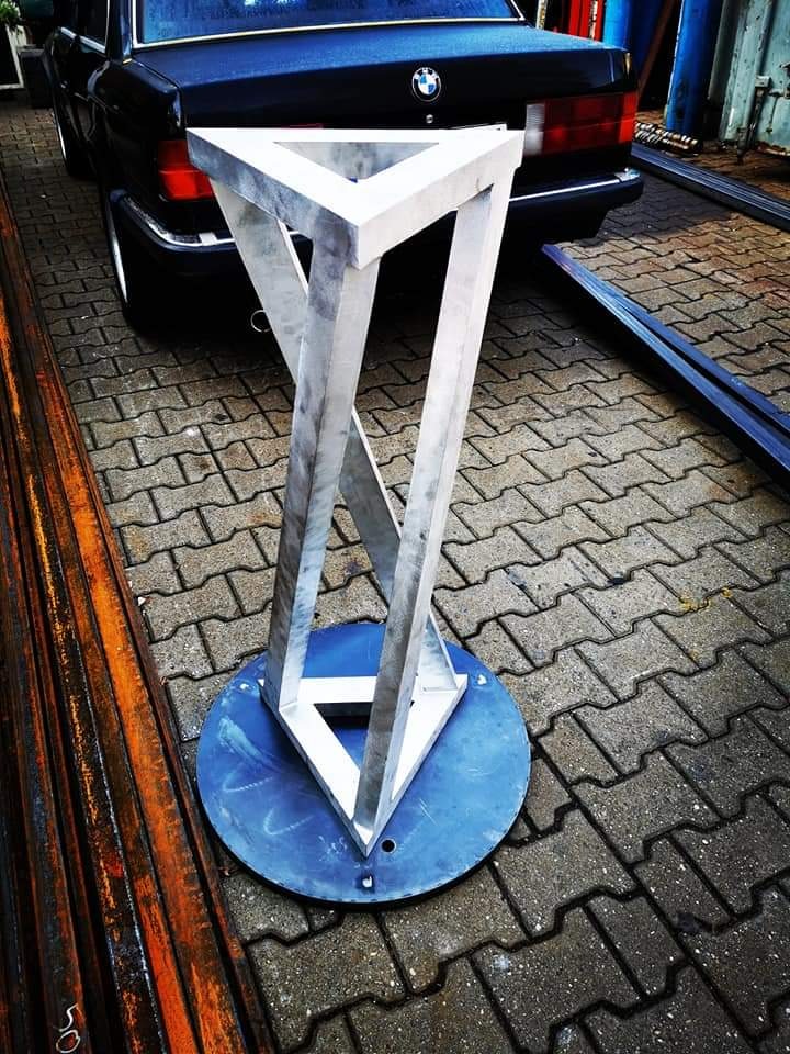 Metalowa konstrukcja o kształcie trójkąta Penrose, spawana z profili, na okrągłej podstawie, prezentowana na kostce brukowej z elementami konstrukcyjnymi i samochodem BMW w tle.