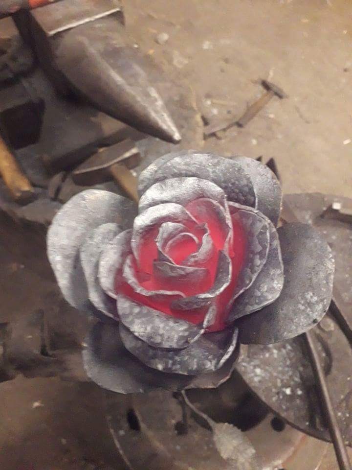 Ręcznie kuta, metalowa róża z czerwonym wnętrzem, leżąca na brudnym stole warsztatowym obok kowadła i narzędzi.