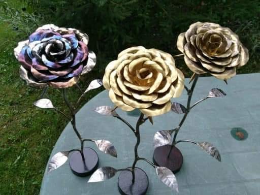 Trzy metalowe róże na ciemnych podstawkach: jedna wielobarwna, druga złota, trzecia miedziana, z metalowymi liśćmi, na tle zieleni.