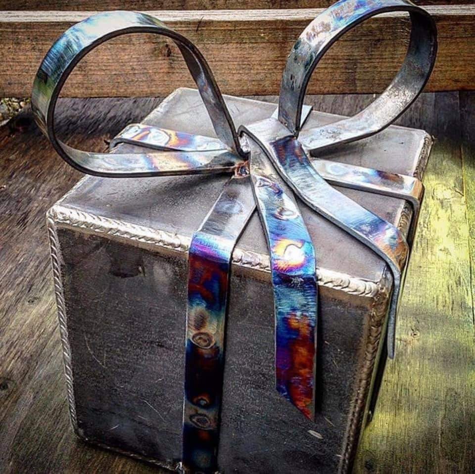 Metalowy prezent z ręcznie spawaną wstążką, widoczne ślady obróbki termicznej i spawania, umieszczony na drewnianym tle.