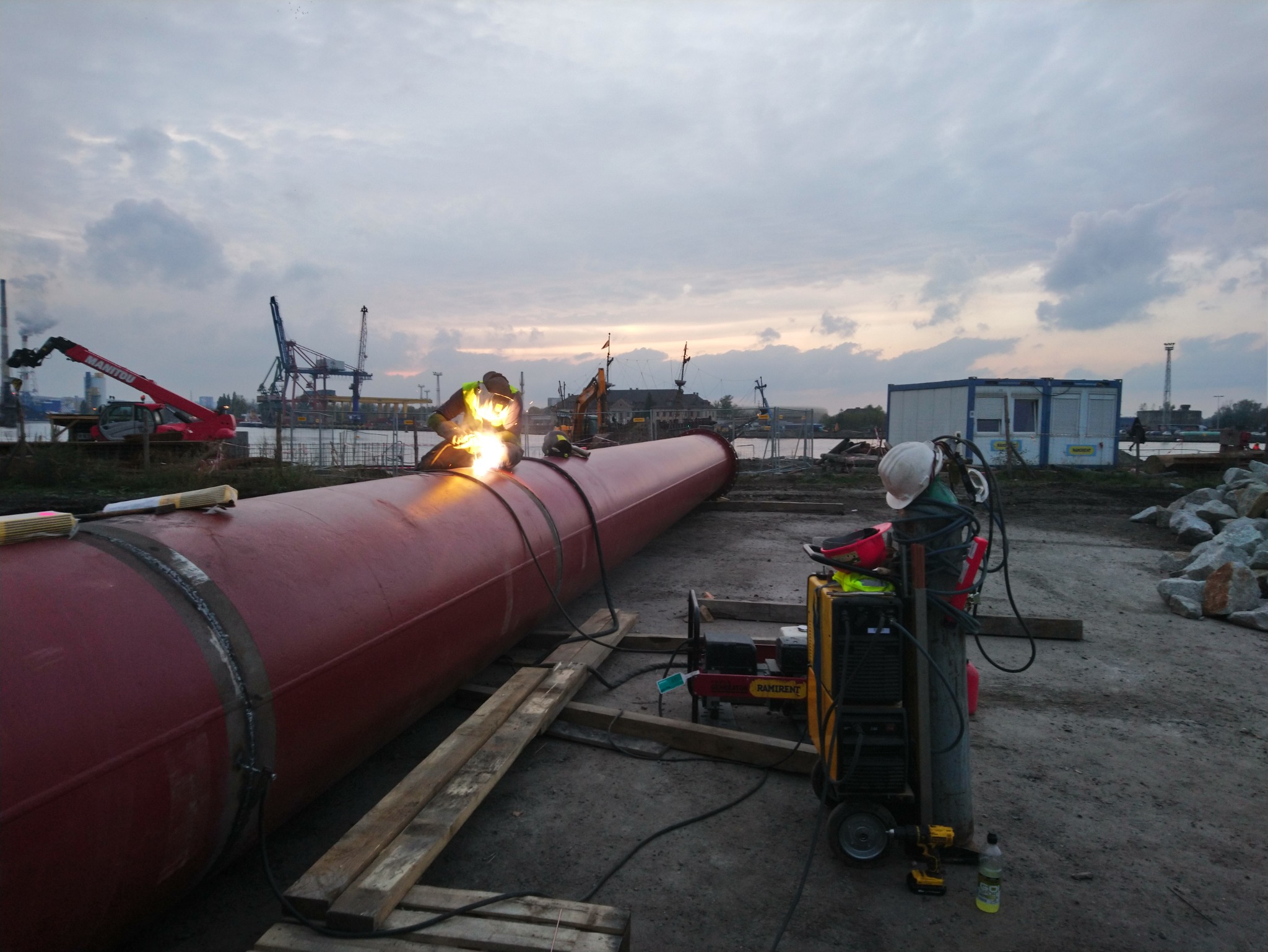 Pracownik w kombinezonie spawalniczym łączy grubą, czerwoną rurę na placu budowy, iskry i jasne światło spawania rozświetlają otoczenie, w tle dźwigi portowe i pochmurne niebo.