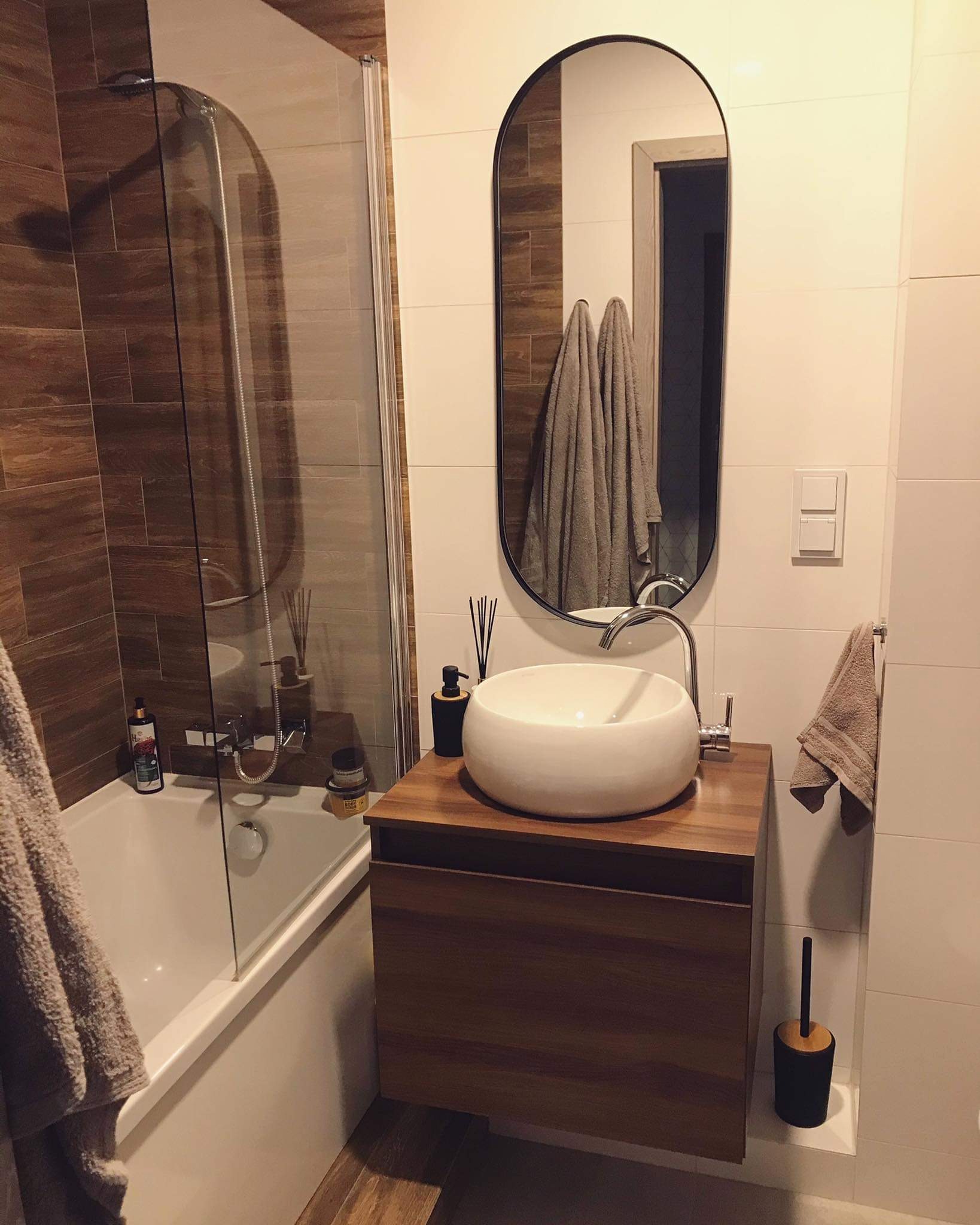 Nowoczesna łazienka z owalnym lustrem, umywalką na drewnianej szafce, kabiną prysznicową i brązowymi oraz białymi płytkami.